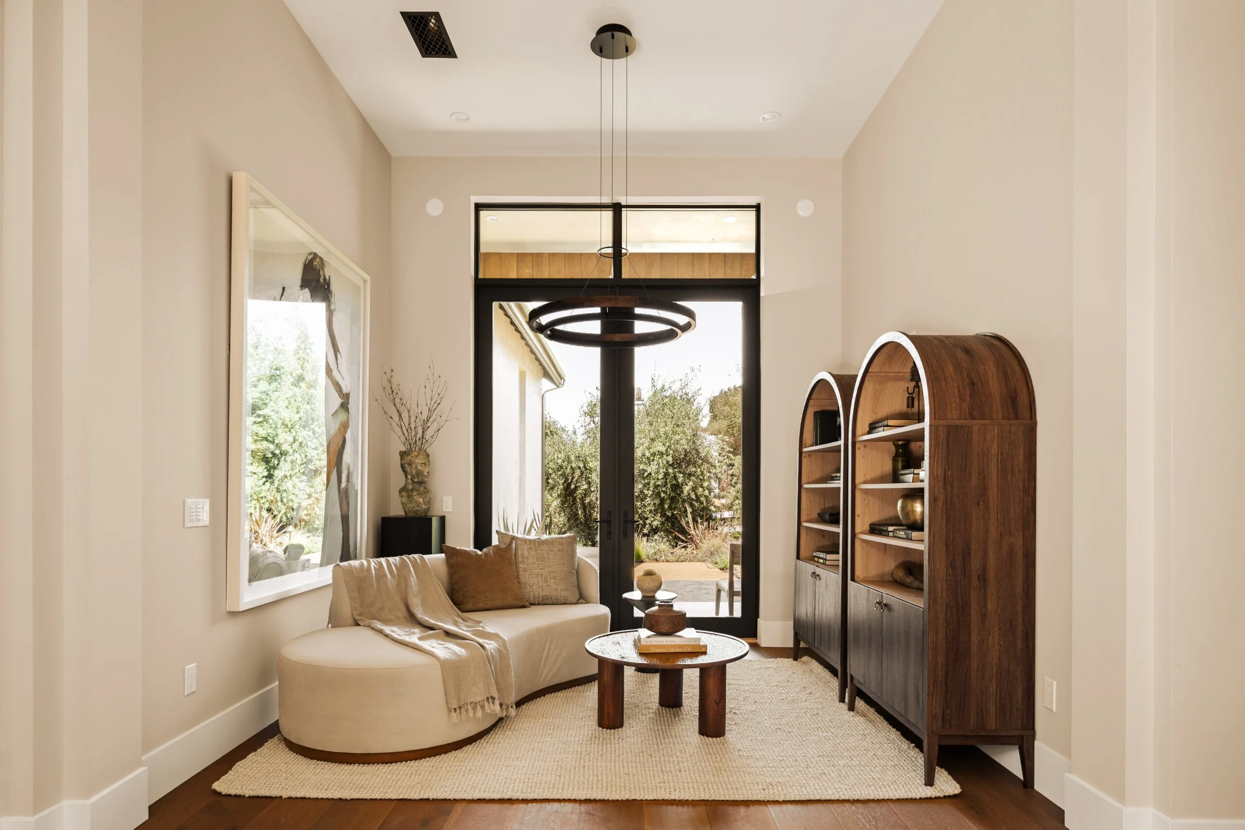 Living room with white curved sofa, beige pillows, a beige throw blanket, a textured rug, a round wooden coffee table, tall wooden bookshelves with decor, a large window with a door opening to outdoor greenery, and a modern circular black chandelier.