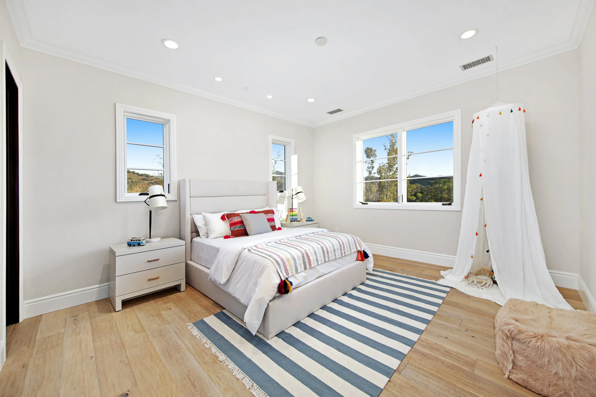 A bright bedroom with white walls, a white bed with colorful pillows, and natural wood flooring. There are three windows showing a scenic outdoor view. A striped rug is placed under the bed, and a small white nightstand with a lamp and small objects 