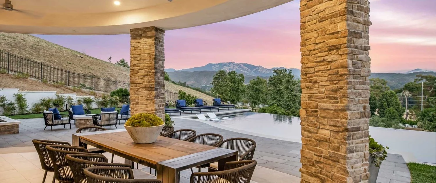 Outdoor patio with stone columns, wooden dining table with chairs, pool area with lounge chairs, mountains and trees in the background, at sunset.