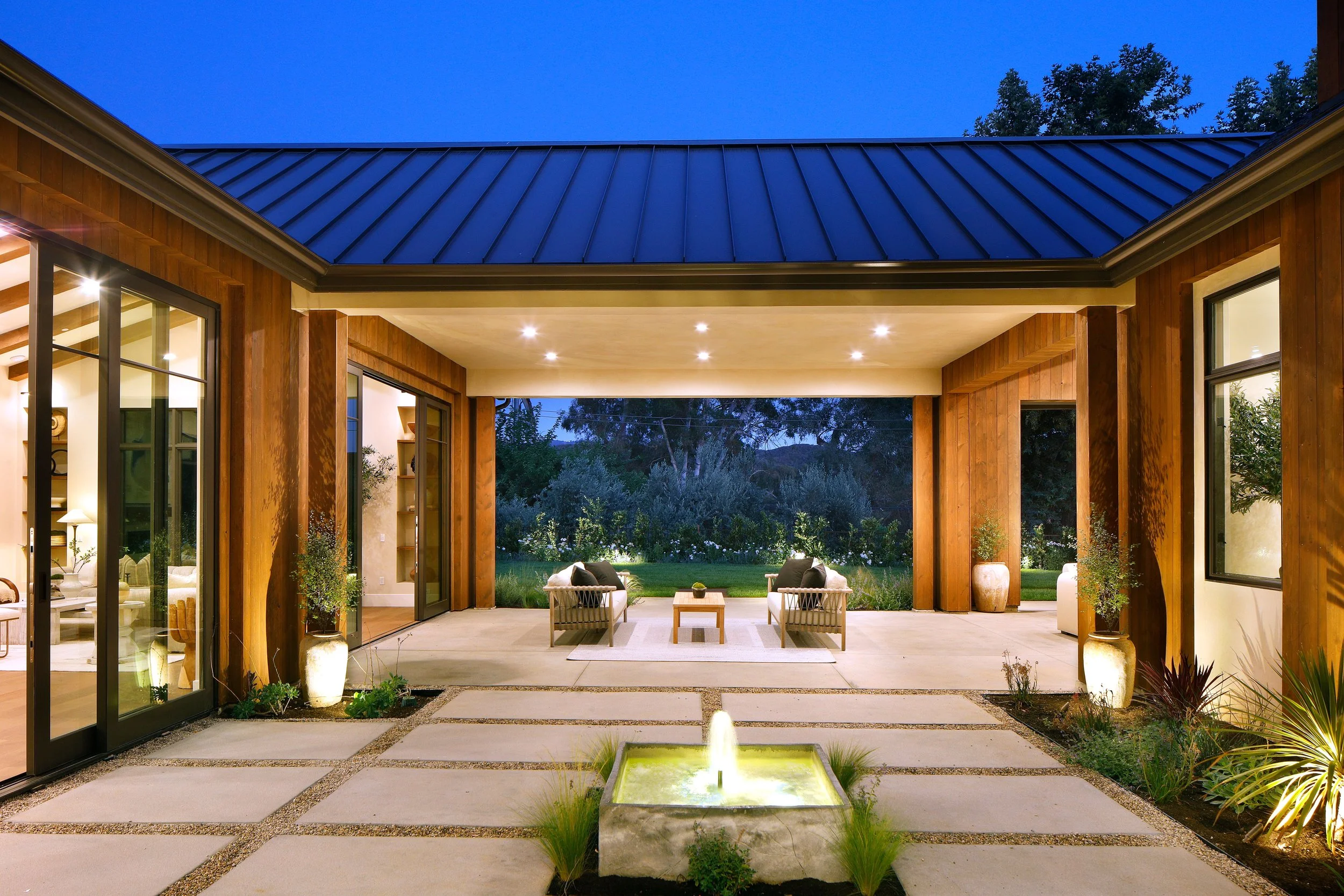 Modern outdoor patio area with seating, decorative potted plants, a water fountain, and view of a garden at dusk.