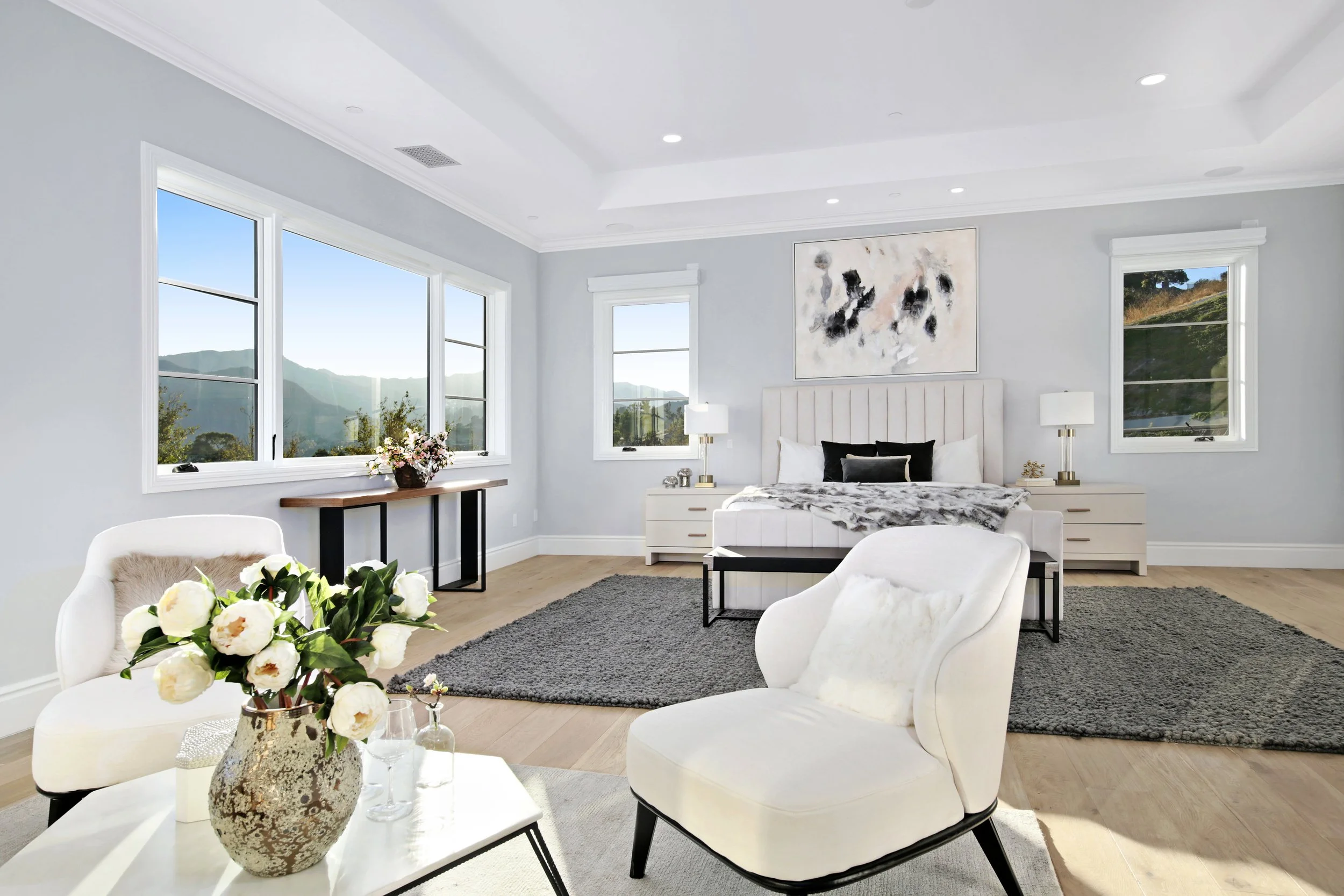 Bright bedroom with large windows, white walls, a bed with white bedding and black pillows, a gray area rug, and modern furniture including white chairs, side tables with lamps, and a wooden console table with a flower arrangement.