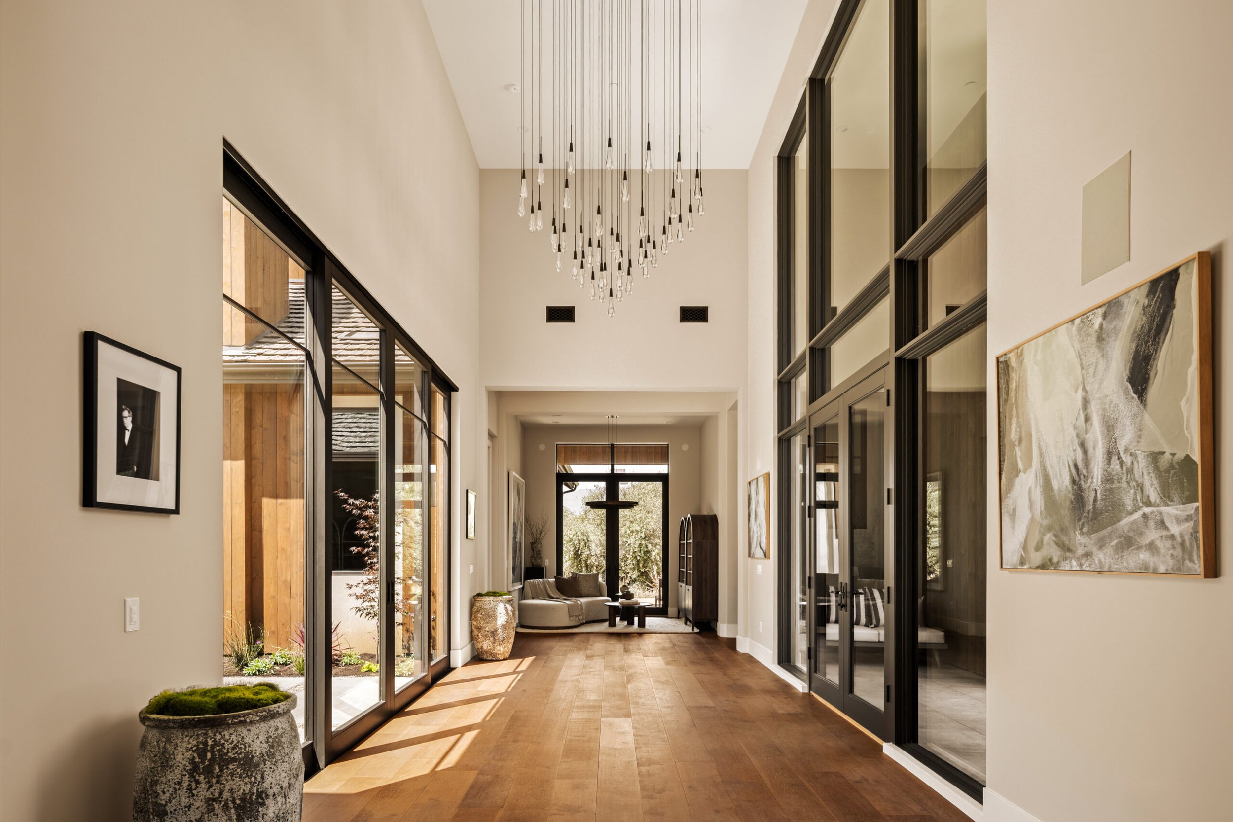 Modern interior hallway with high ceiling, large black framed glass doors, wooden flooring, and contemporary art on beige walls, leading to a sitting area with a curved sofa and large window with views of greenery.