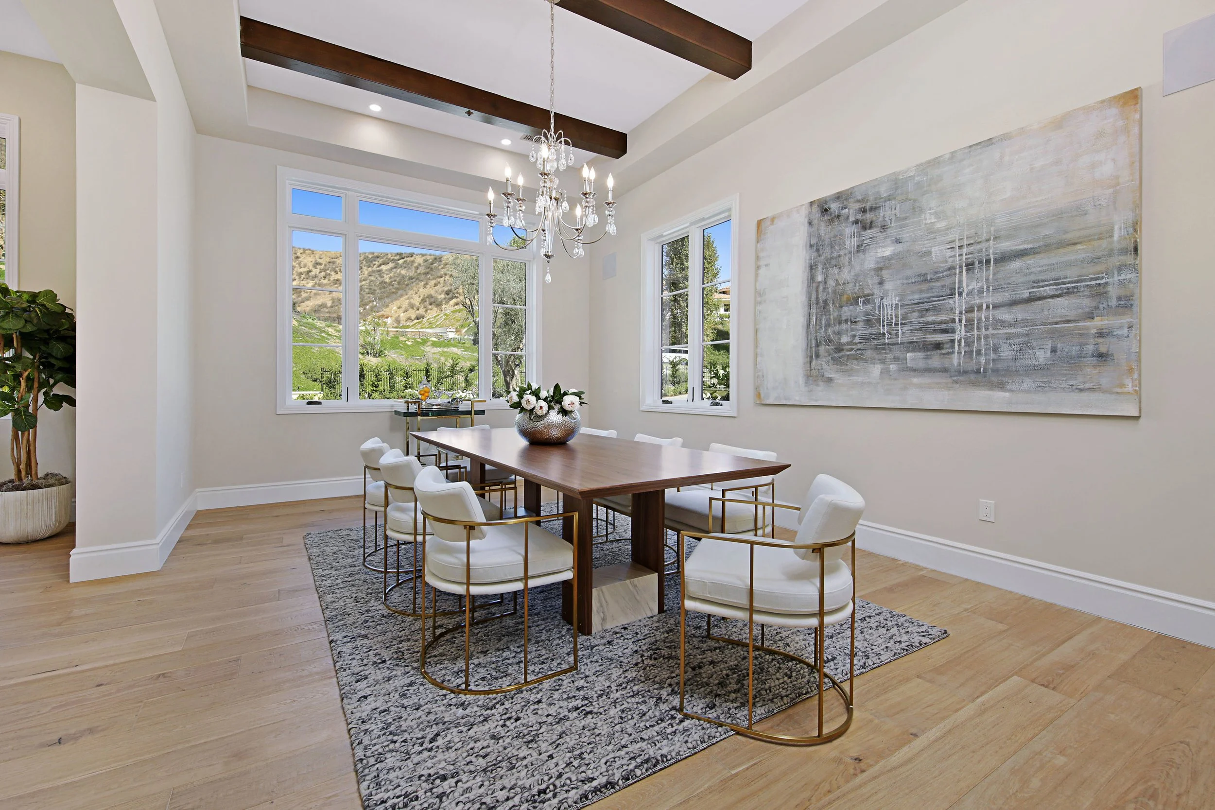 Bright dining room with large windows overlooking greenery and hills, wooden dining table with eight white cushioned chairs with gold frames, chandelier overhead, large abstract painting on wall, plant in corner, rug under table.