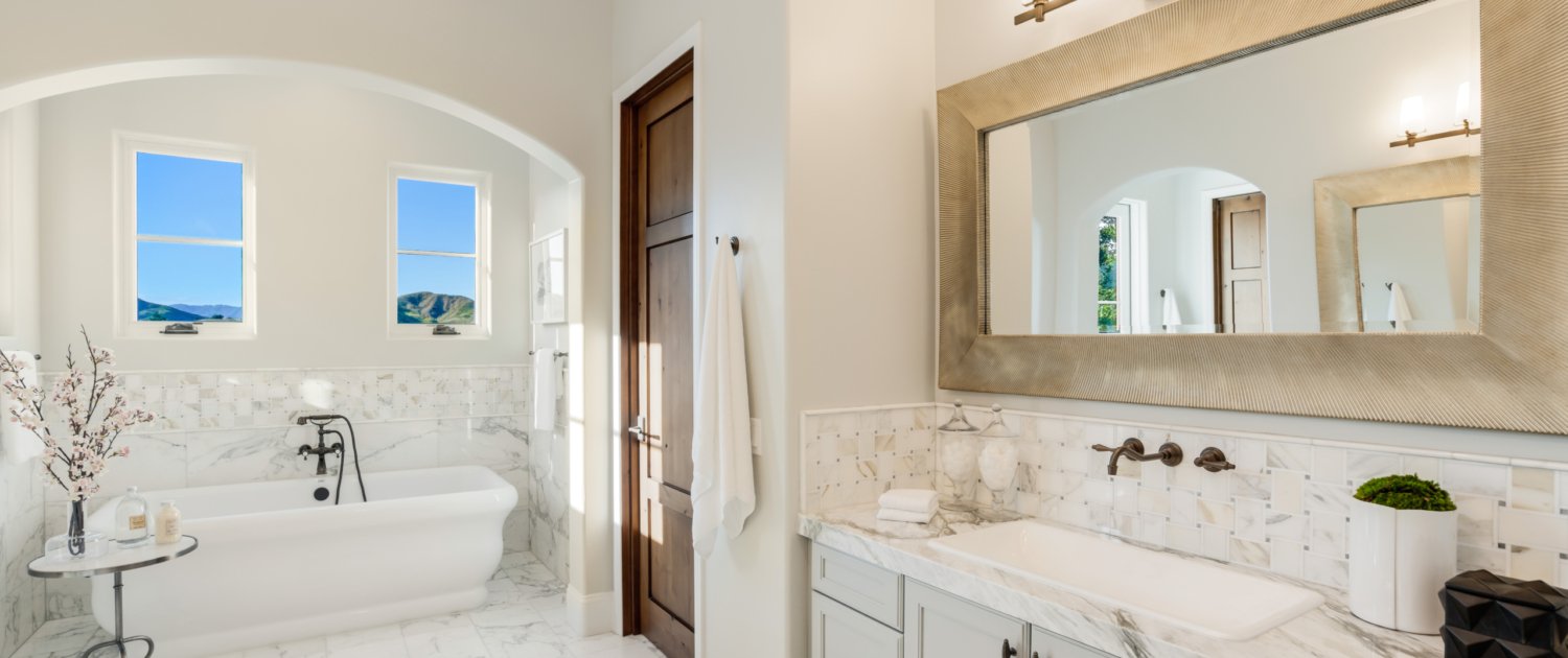 A bright and airy bathroom with a white bathtub, two windows showing a mountain landscape, a marble countertop, and a large rectangular mirror.