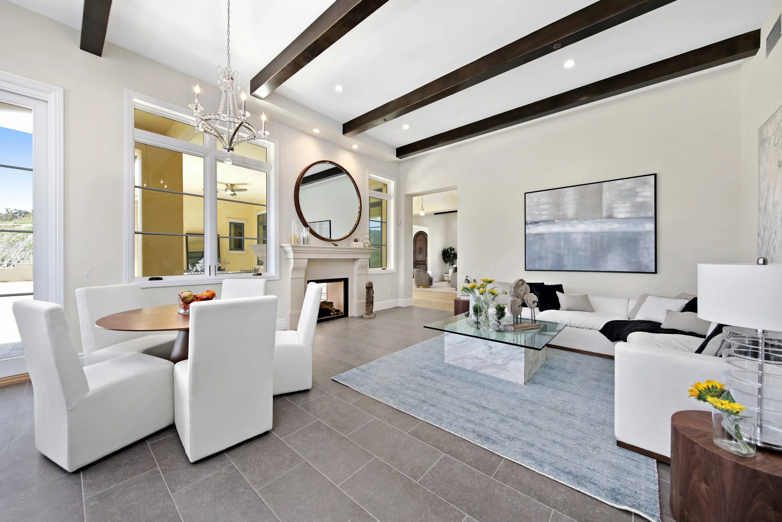 Spacious living room with modern furniture, white sectional sofa, glass coffee table, flat-screen TV, fireplace, and dining area with white chairs and wooden table. Large windows with view outside, dark wood beams on ceiling, and neutral color palett