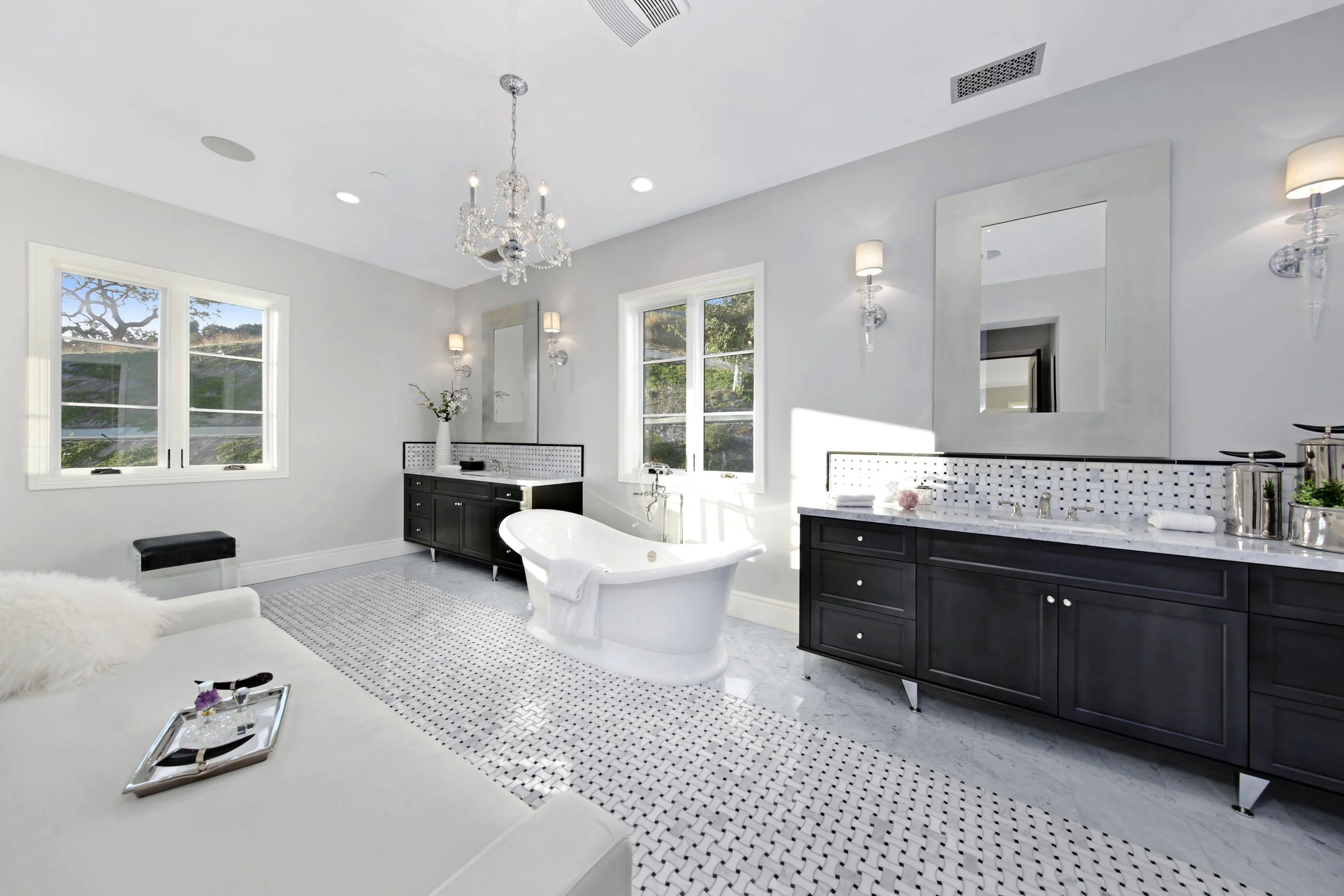 Bright bathroom with two black vanities, a white clawfoot bathtub, a patterned rug, large windows, and elegant wall-mounted lights.