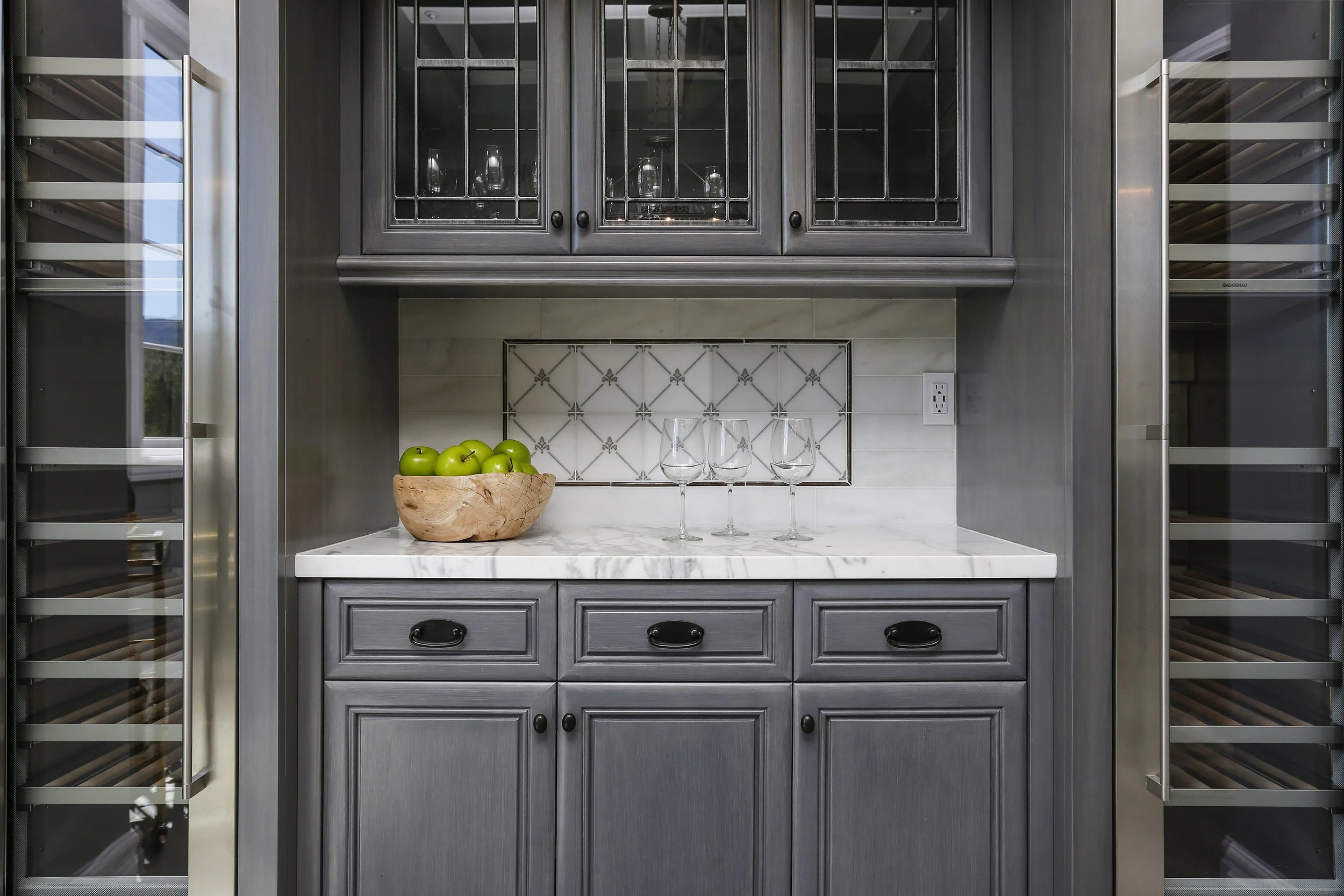 A built-in wine storage and bar area with gray cabinets, a marble countertop, a bowl of green apples, three wine glasses, and a decorative tiled backsplash.