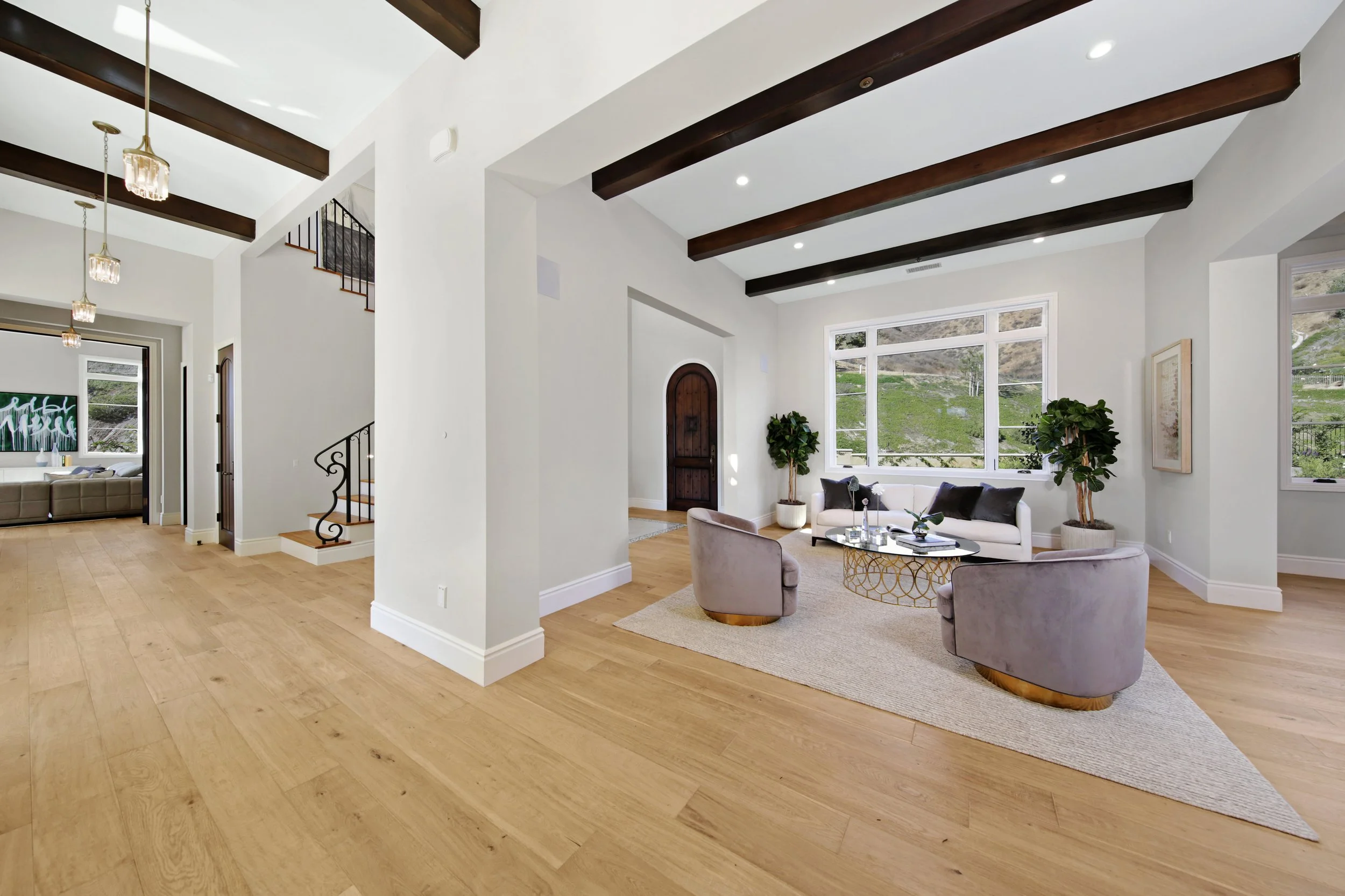 Living room with white walls, hardwood floors, large windows, and dark wooden ceiling beams. Contains a white sofa, two gray armchairs, a gold coffee table, and potted plants.