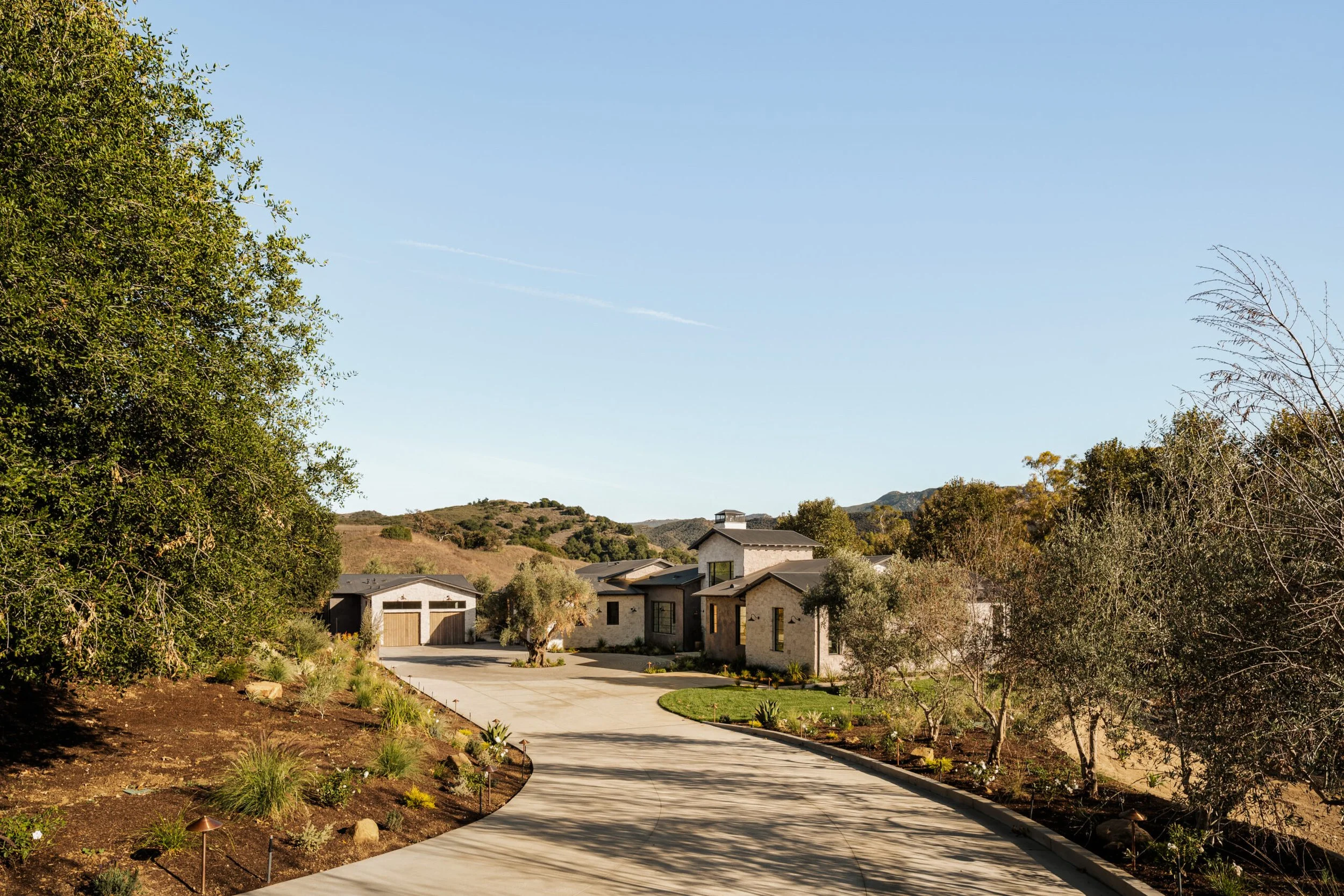 A driveway leading to a house in a rural, hilly area with trees and landscaping on either side.