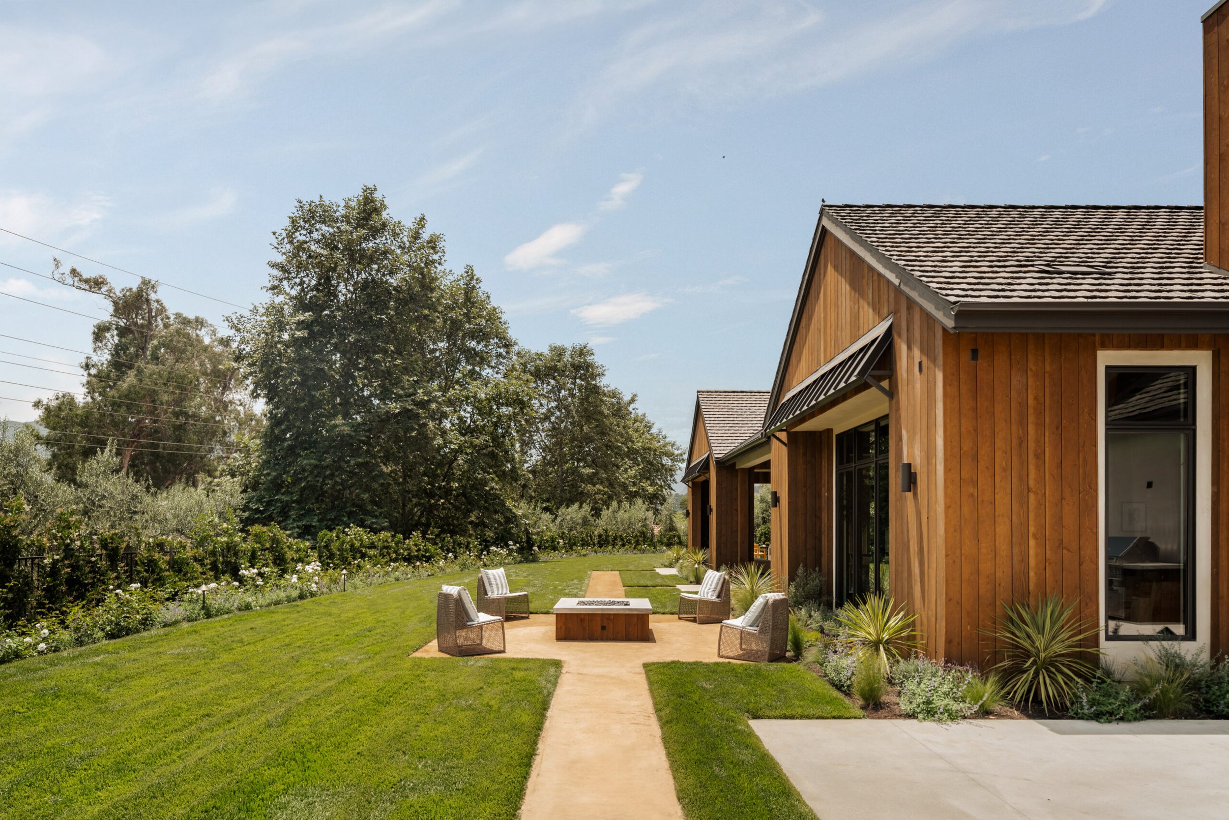 Exterior view of modern wooden house with a backyard patio with chairs and fire pit, on a sunny day.