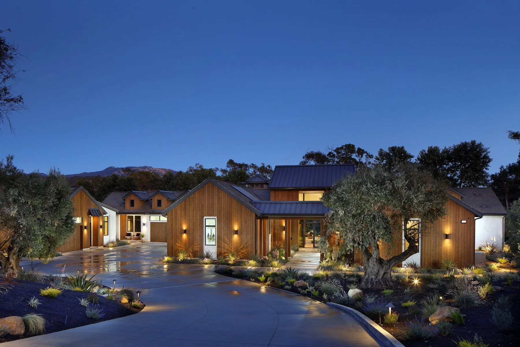 Modern house with wooden exterior and landscaped front yard at dusk, with trees, pathway, and outdoor lighting.