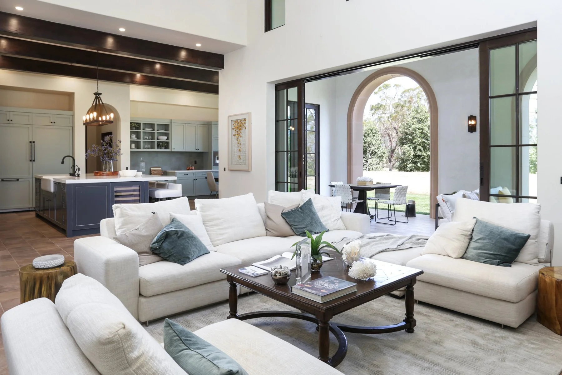 A spacious, modern living room with white sofas, dark wooden coffee table, and blue throw pillows. Open kitchen with a large island, light-colored cabinets, and a dining area with outdoor view through large glass doors.