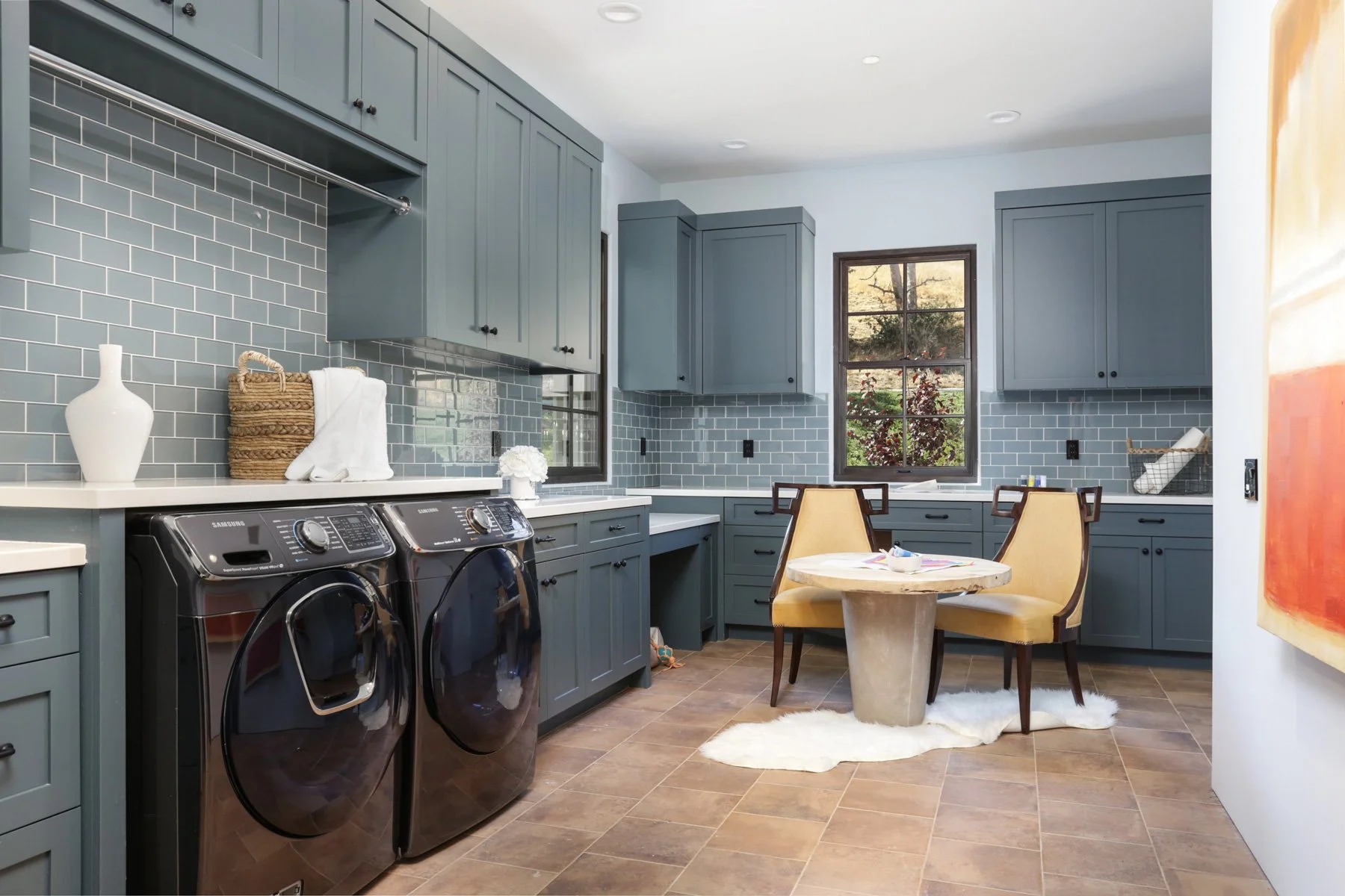 A laundry room with dark gray cabinets, gray subway tile backsplash, washer and dryer, and a small round table with two chairs.