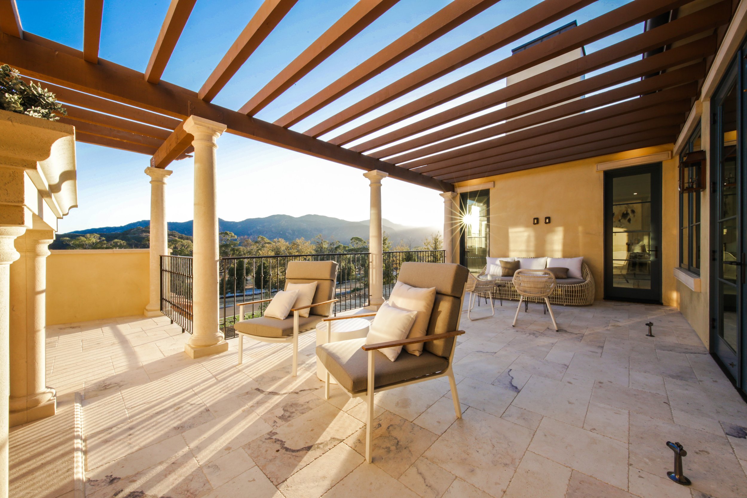 Sunlit outdoor balcony with beige tile flooring, two cushioned chairs with armrests, a wicker sofa with cushions, a small glass table, and a mountain view in the background. The balcony has a wooden pergola and columns, with the sun shining through.