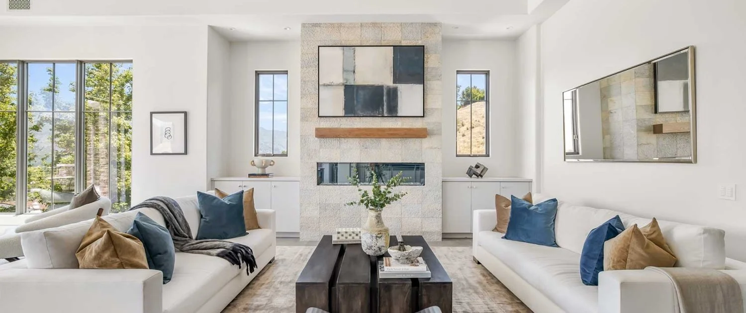 Modern living room with white walls, large windows, two white sofas with beige and dark blue pillows, a black coffee table with a vase and books, a large abstract painting above the fireplace, and a flat-screen TV on the right wall.