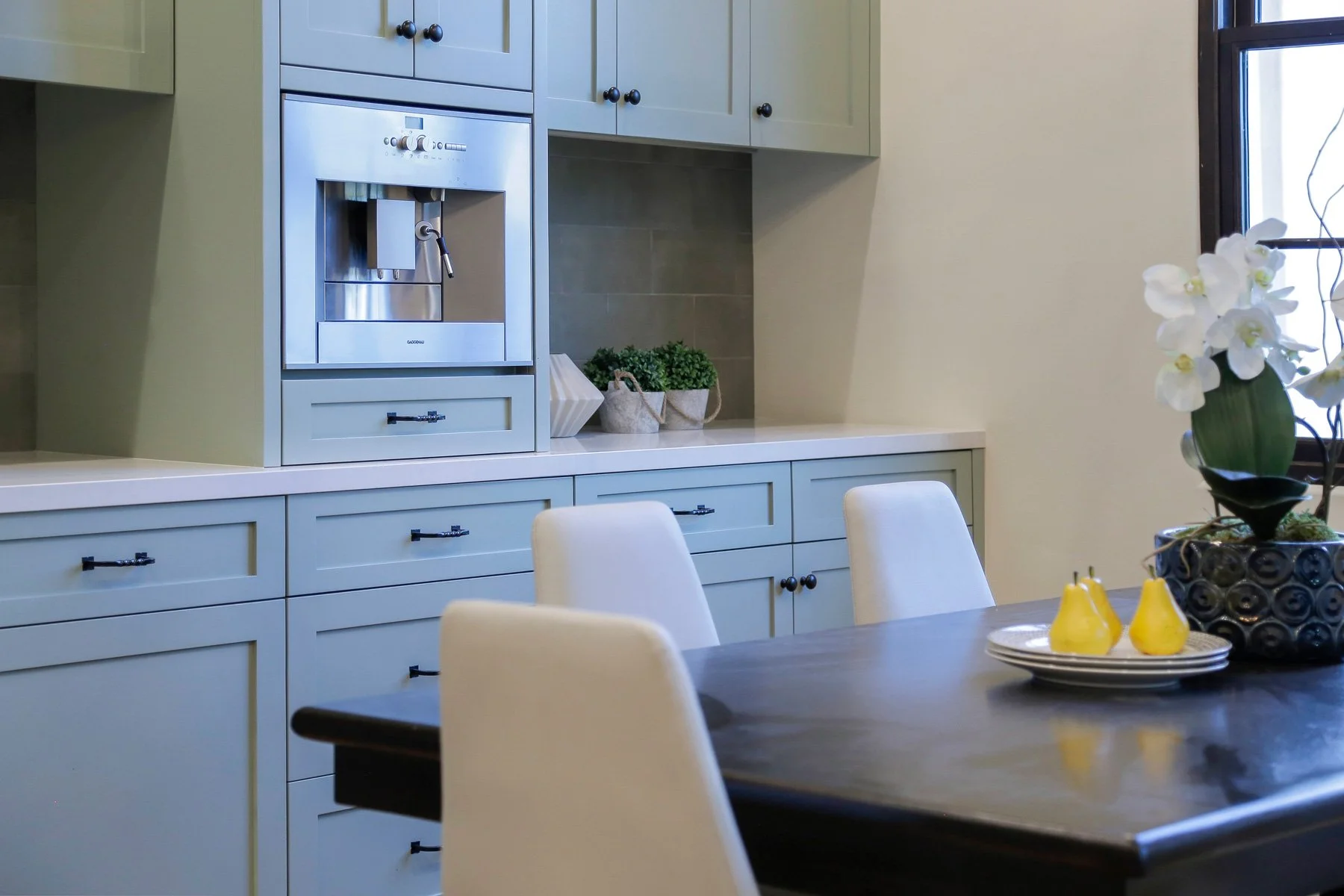 A modern kitchen with light green cabinets and a built-in stainless steel coffee maker. There is a dark wood dining table with white upholstered chairs, a flower arrangement with white orchids, and a window in the background.
