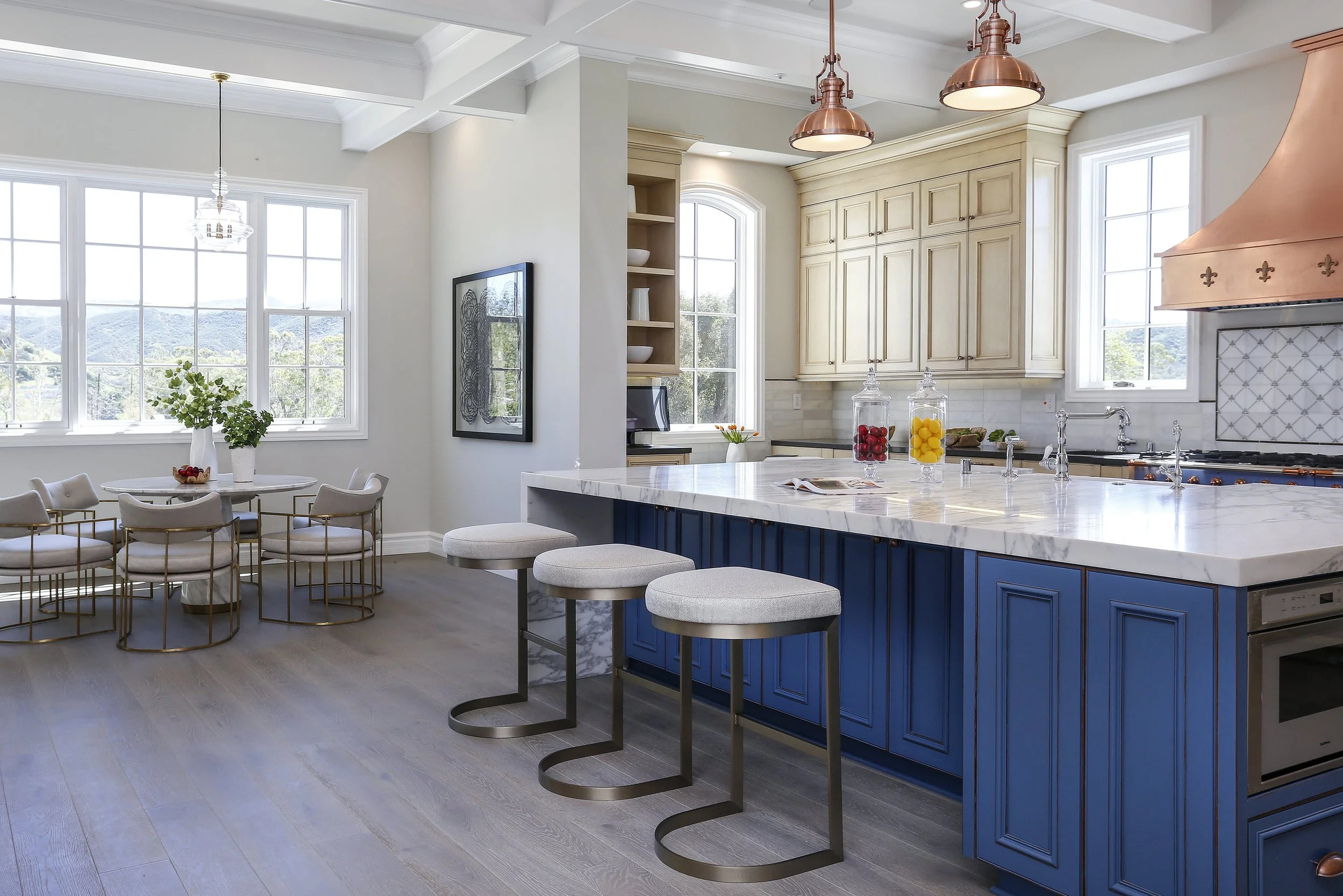 Modern kitchen with blue cabinets, white marble countertops, copper pendant lights, and large windows showing a scenic view. An adjacent dining area has a round table with eight upholstered chairs, a chandelier, and greenery in a vase.