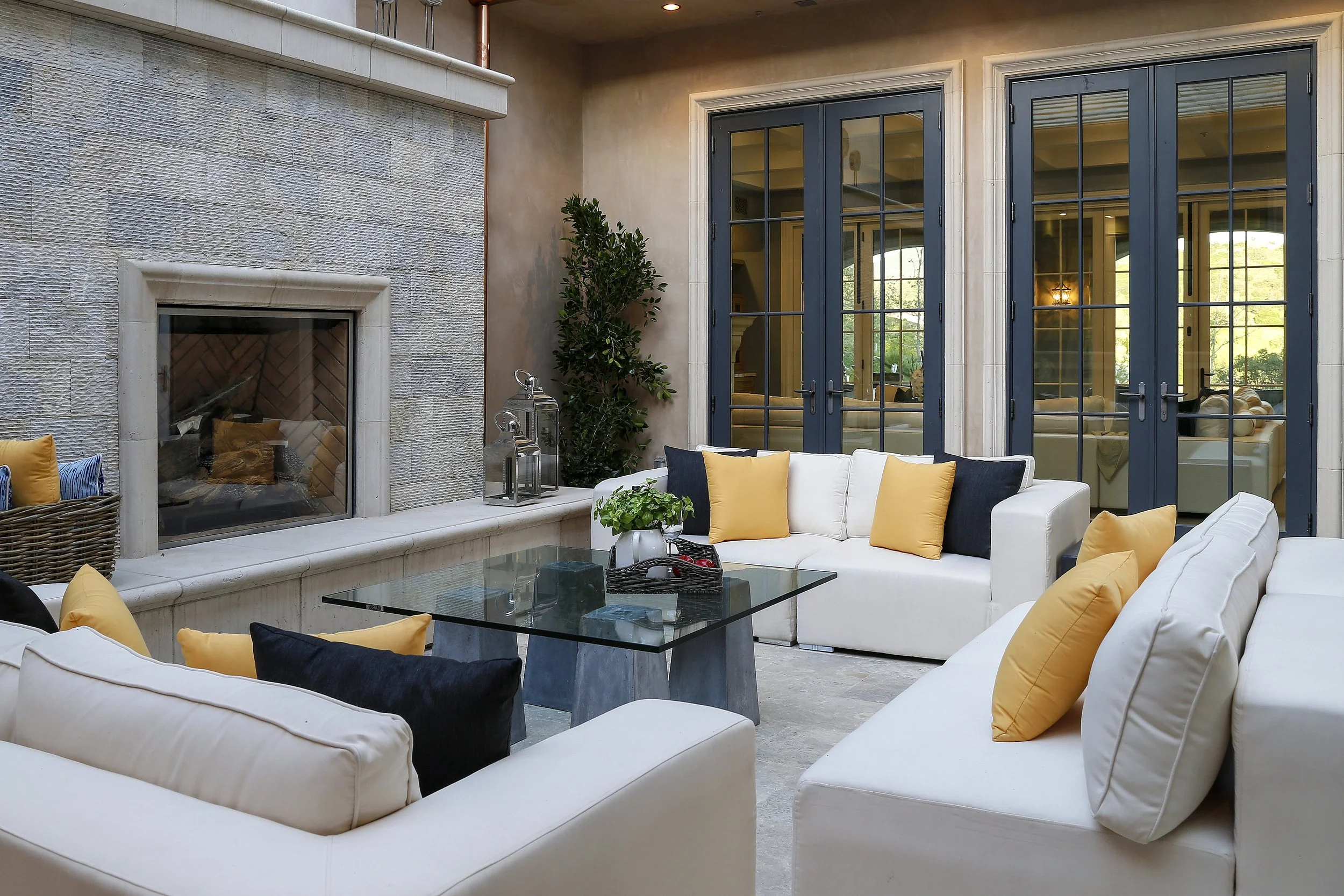 Indoor patio with white sofas decorated with yellow and black cushions, glass coffee table with a small plant, and a fireplace with a textured stone surround. French doors with blue frames lead outside.