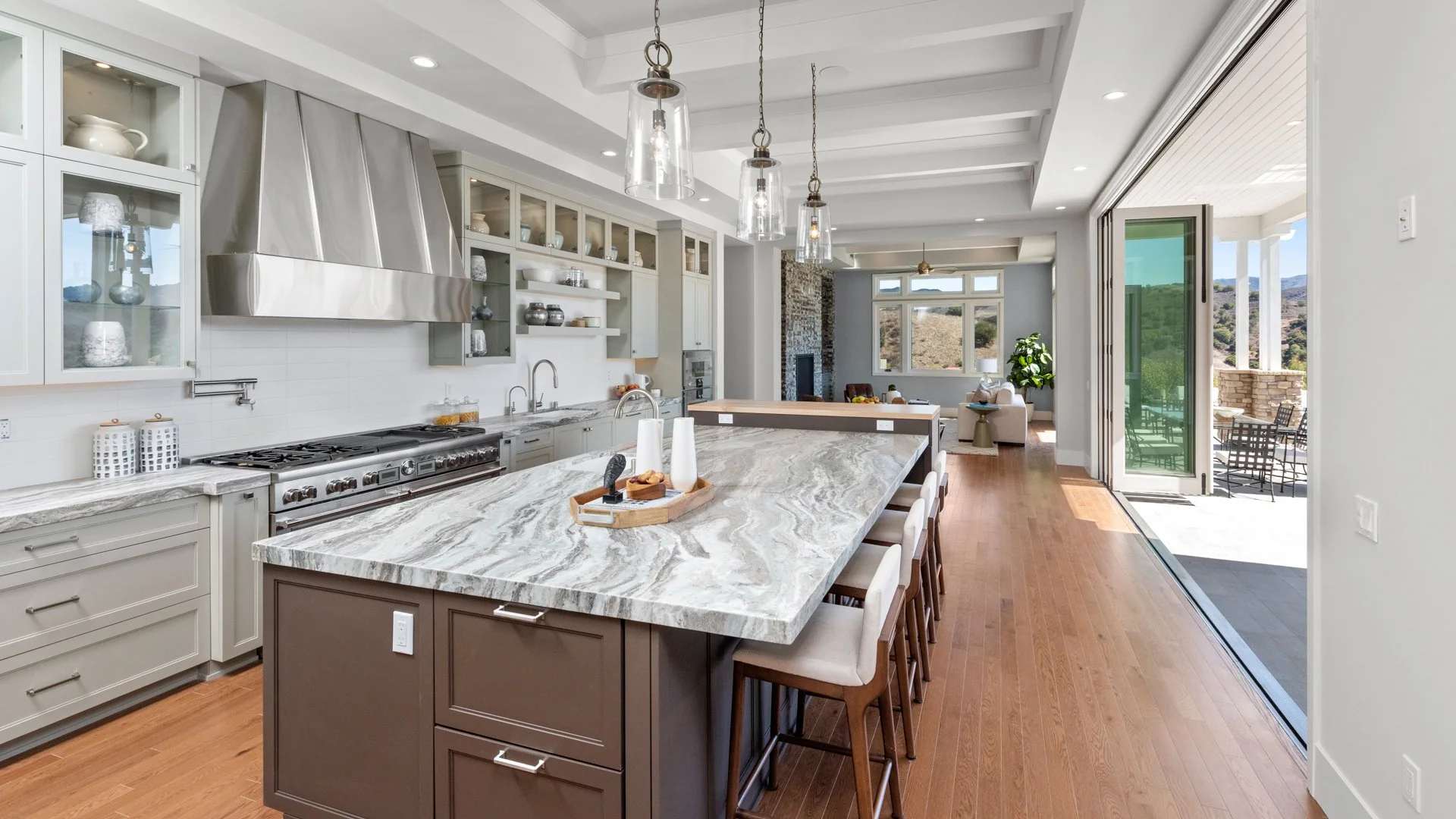 Modern open-concept kitchen with an island, marble countertop, stainless steel appliances, open shelving, pendant lights, and a view into a living area and outdoor patio with chairs.