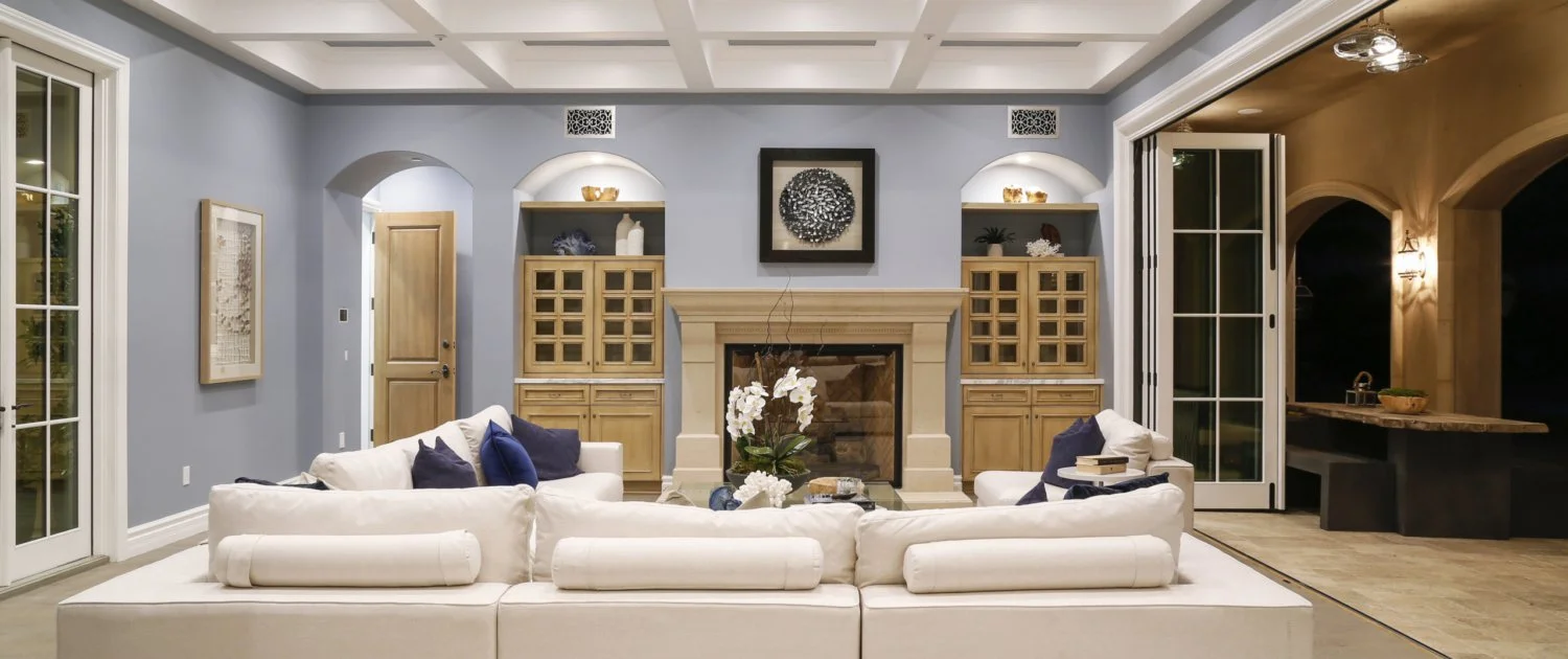 Living room with light blue walls, beige built-in cabinets surrounding a fireplace, a white sectional sofa with dark blue pillows, a glass coffee table with flowers, and sliding glass doors leading outside.