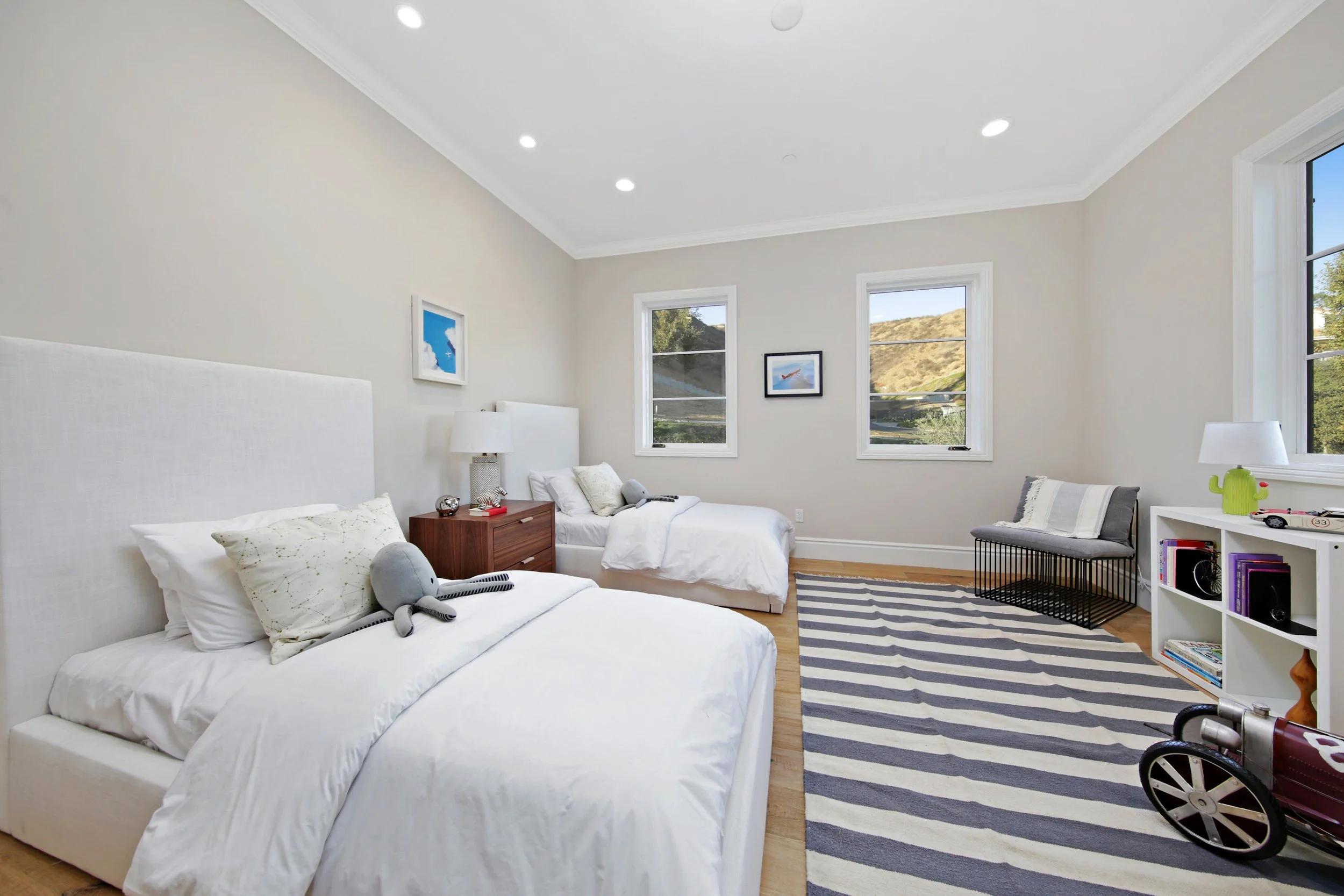 A bedroom with two white beds, a wooden nightstand, a gray bench, a white bookshelf, and three windows showing an outdoor view of hills and trees.
