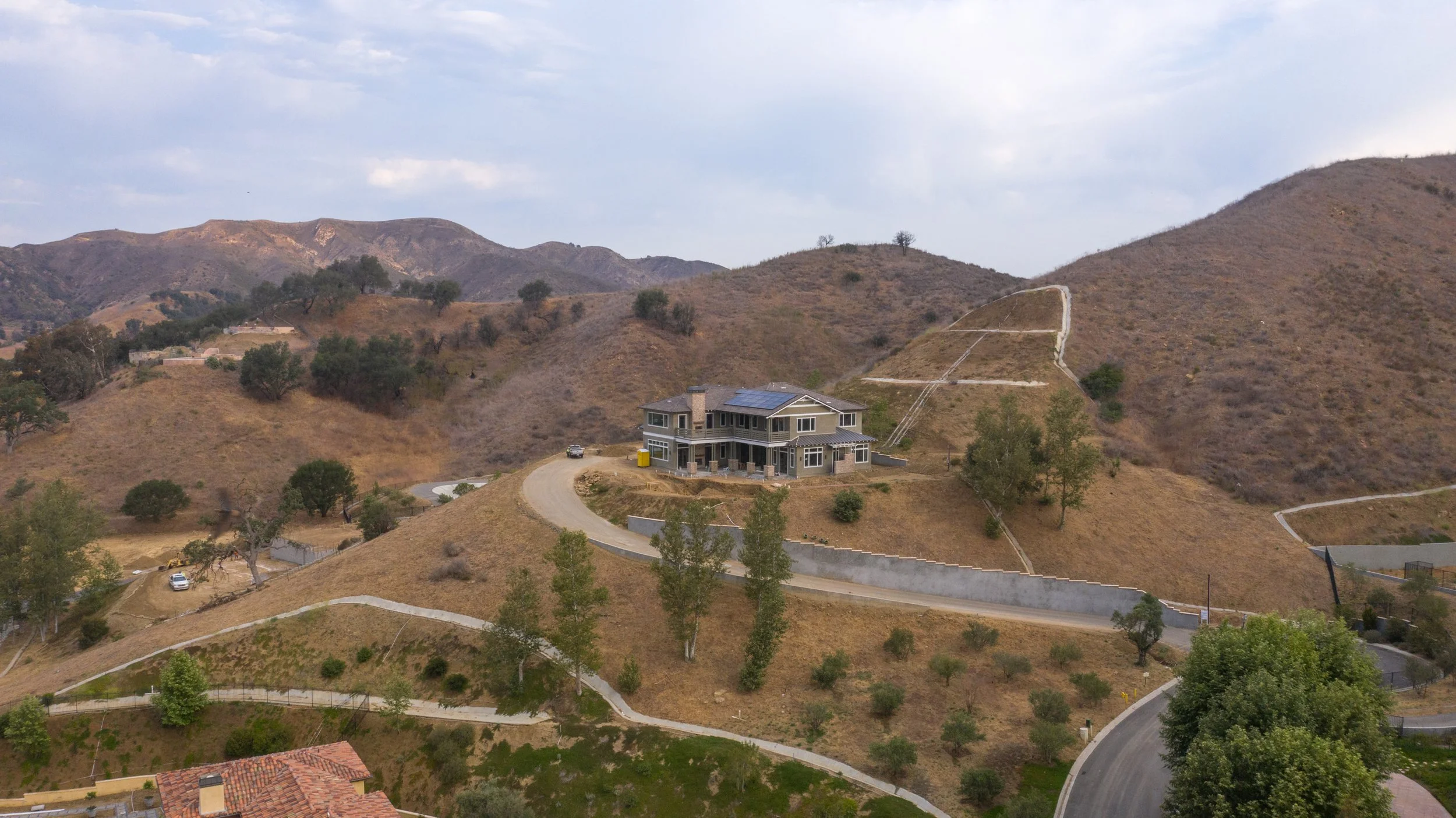 A house situated on a hill with multiple levels, surrounded by winding roads, dry grassy terrain, and a few trees amidst rolling hills and mountains in the background.
