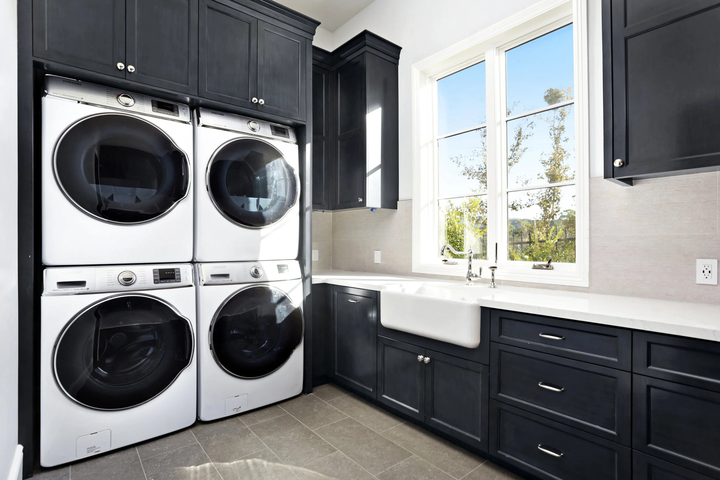 Laundry room with four stacked washer and dryer units, dark blue cabinets, a large white farmhouse sink, and a window with a view of trees outside.