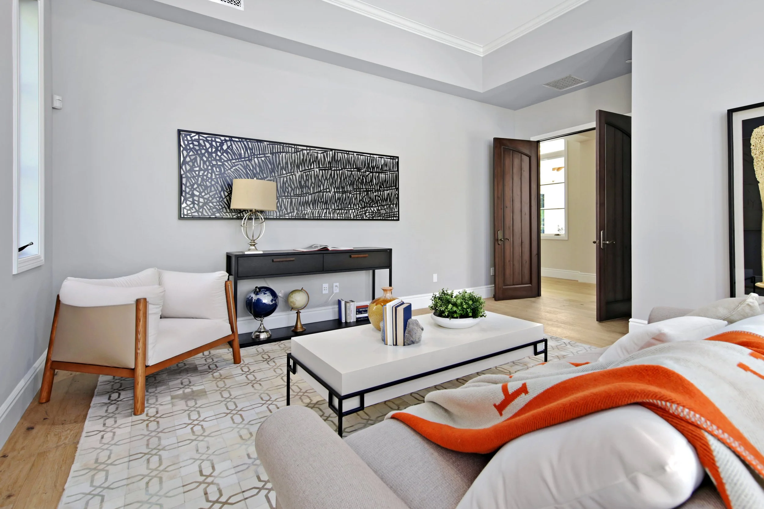 Modern living room with white walls, wooden flooring, a grey rug, a beige sofa with an orange and white blanket, a black console table, a white chair, and a white coffee table with decorative vases and books. Open dark wood double doors lead to anoth