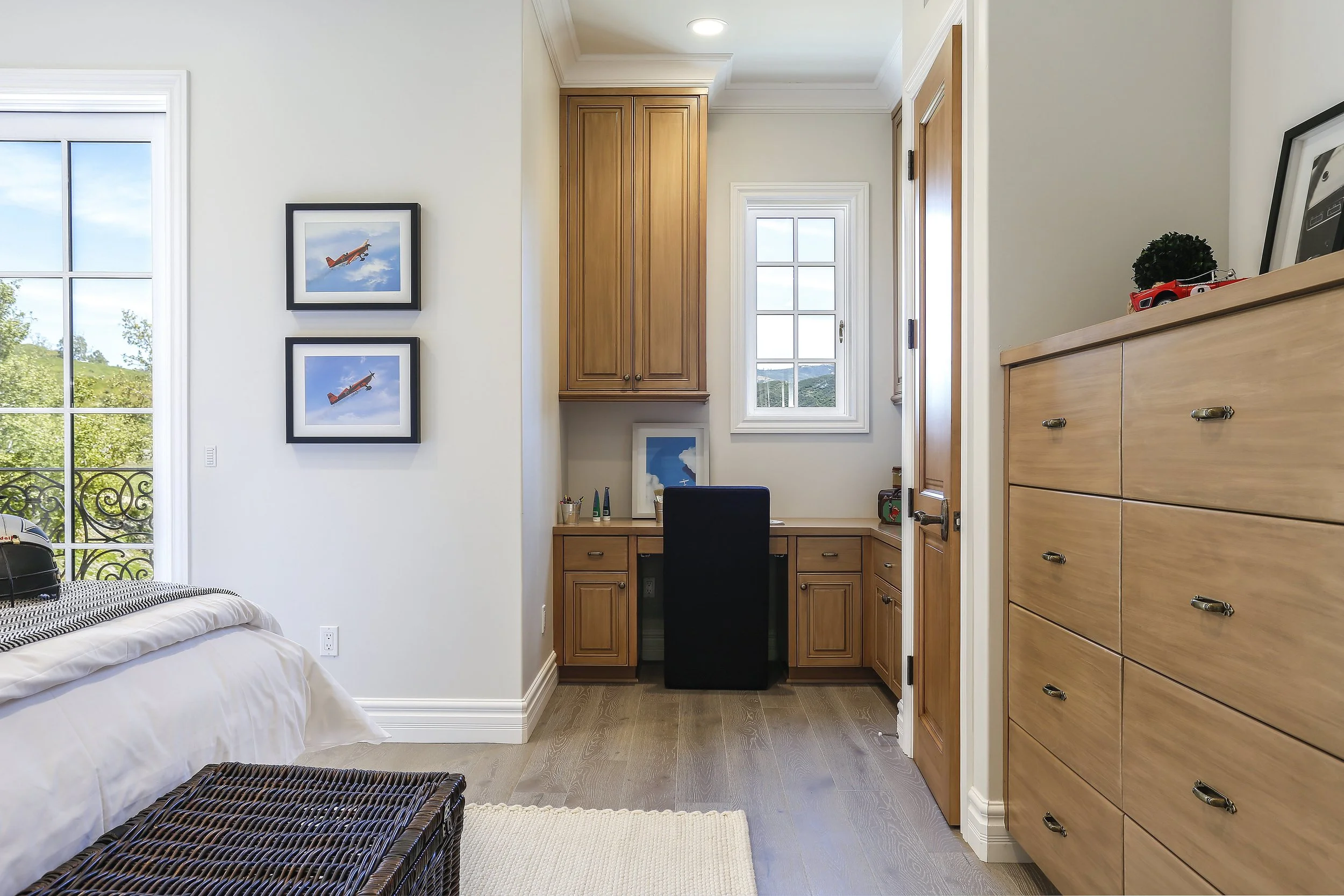 A bedroom corner with a Built-in wooden desk and cabinets, a window, a wooden dresser, and wall art portraying airplanes.