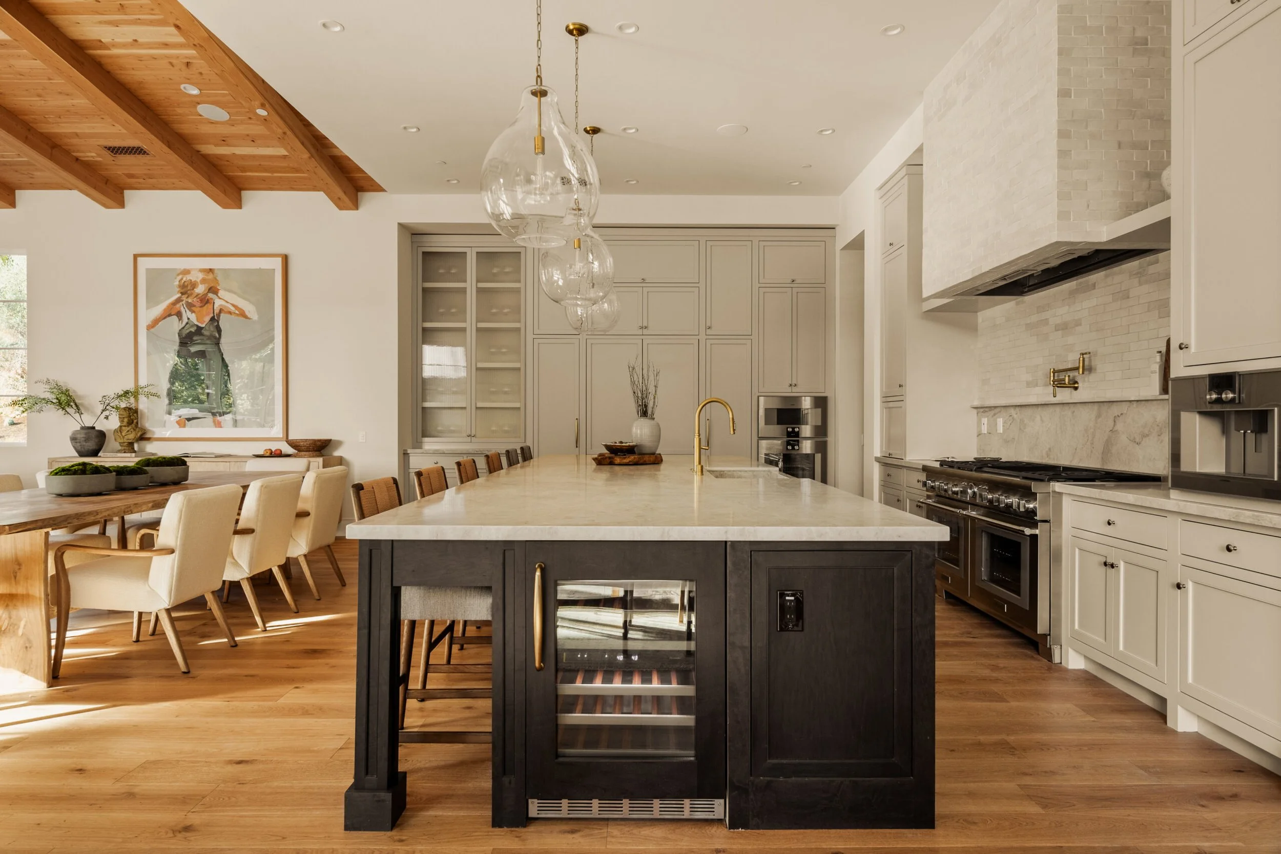 Modern kitchen with a central island, white cabinets, and a large dining table with cream-colored chairs, featuring a wooden ceiling and hardwood floors.