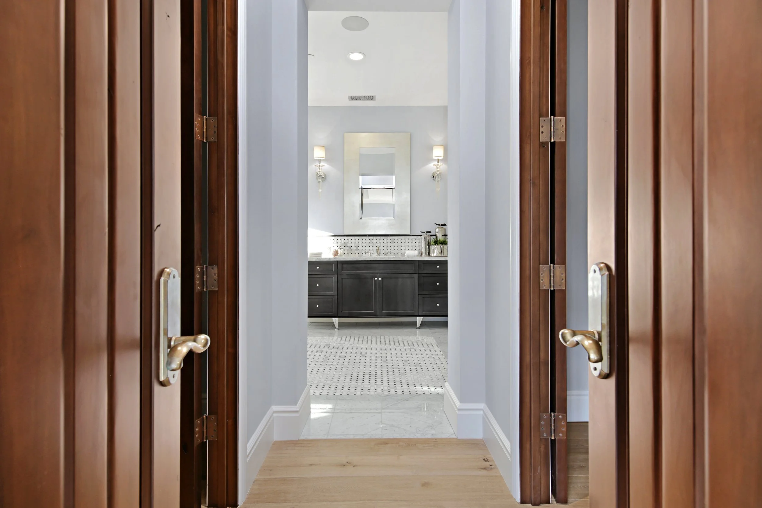 A view through a doorway into a bathroom with a dark vanity, a mirror, and wall sconces, with a window and patterned rug.