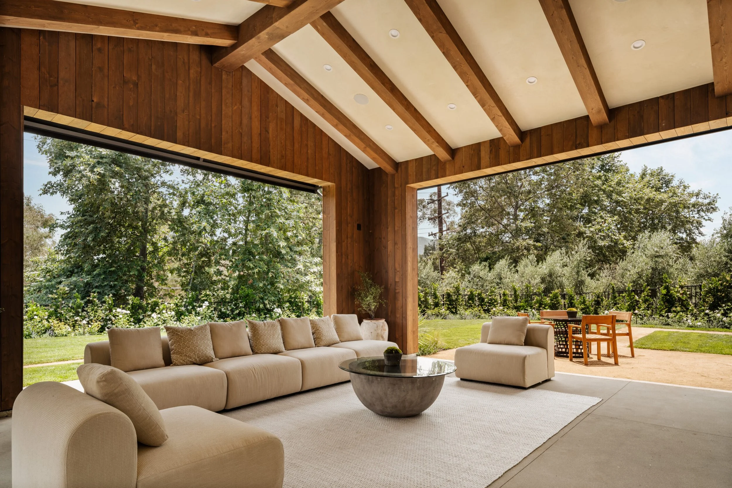 Living room with large beige sectional sofa, a round stone coffee table, and a cream armchair, opening to a lush green backyard with trees and outdoor dining table.