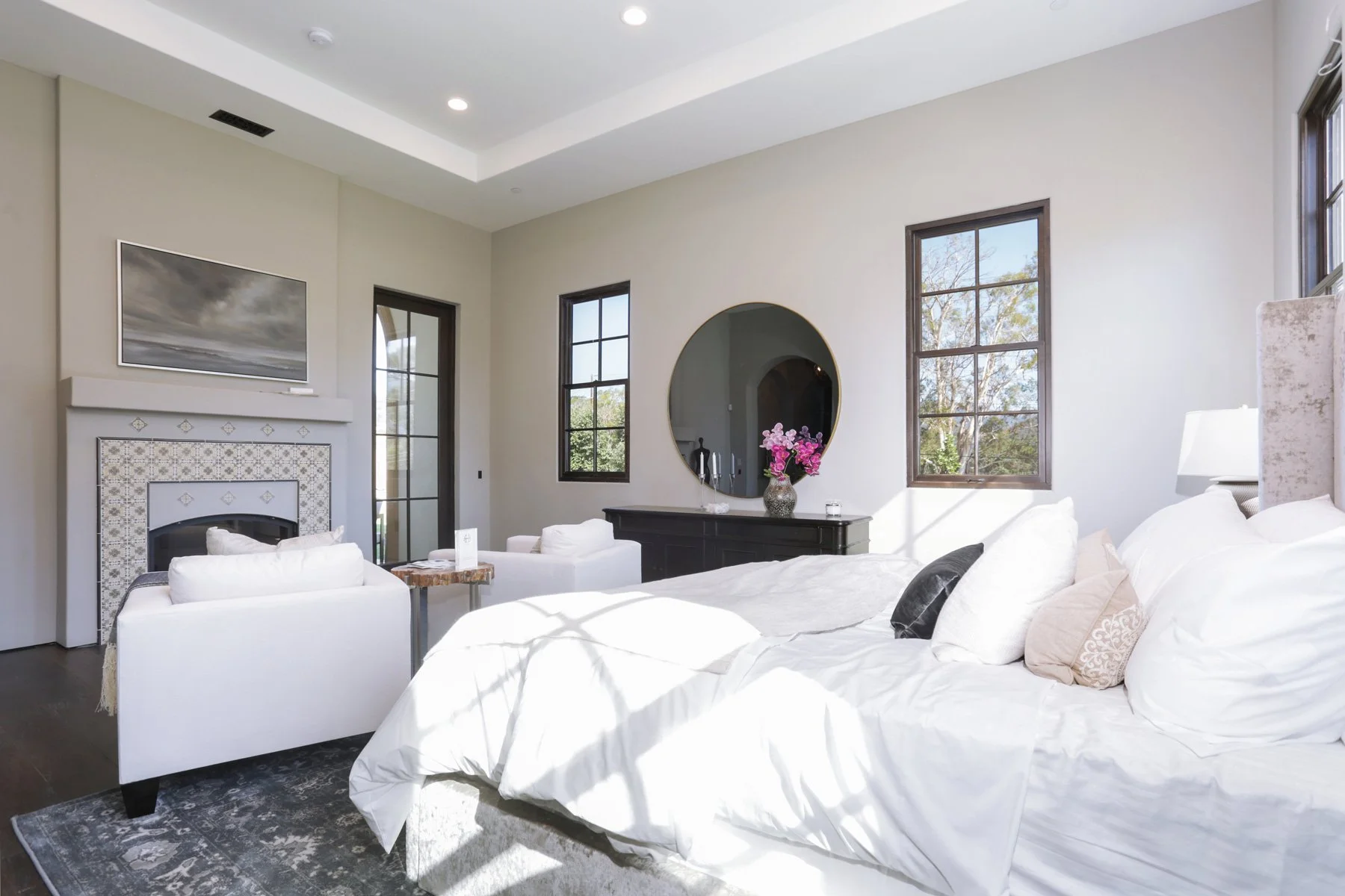 Bright bedroom with white bedding, a large bed, black-framed windows, and a black dresser with a pink flower vase, a round mirror, and natural light coming through the windows.