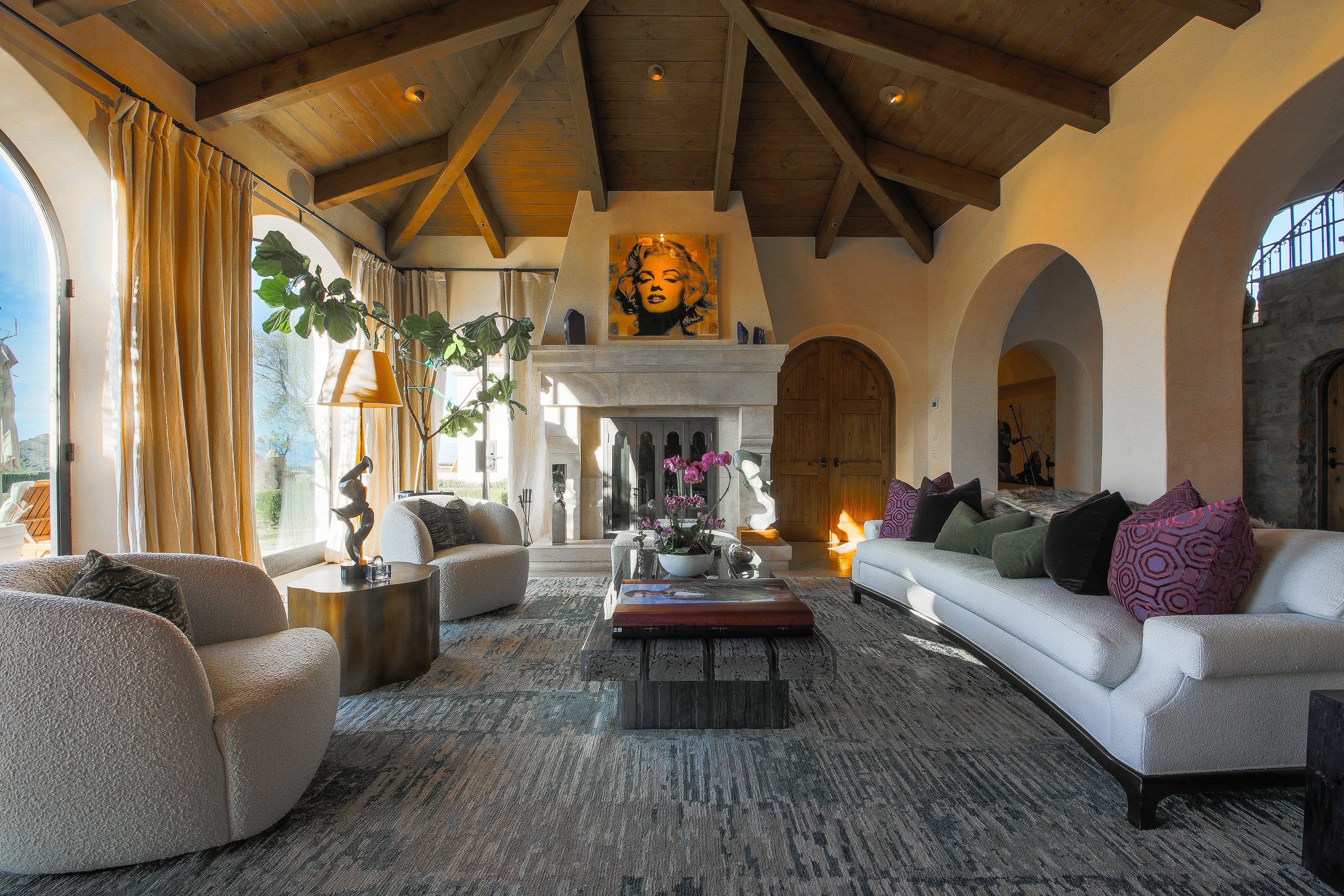 Living room with large windows, beige and white furniture, a fireplace with a Marilyn Monroe artwork above it, and a wooden ceiling with exposed beams.