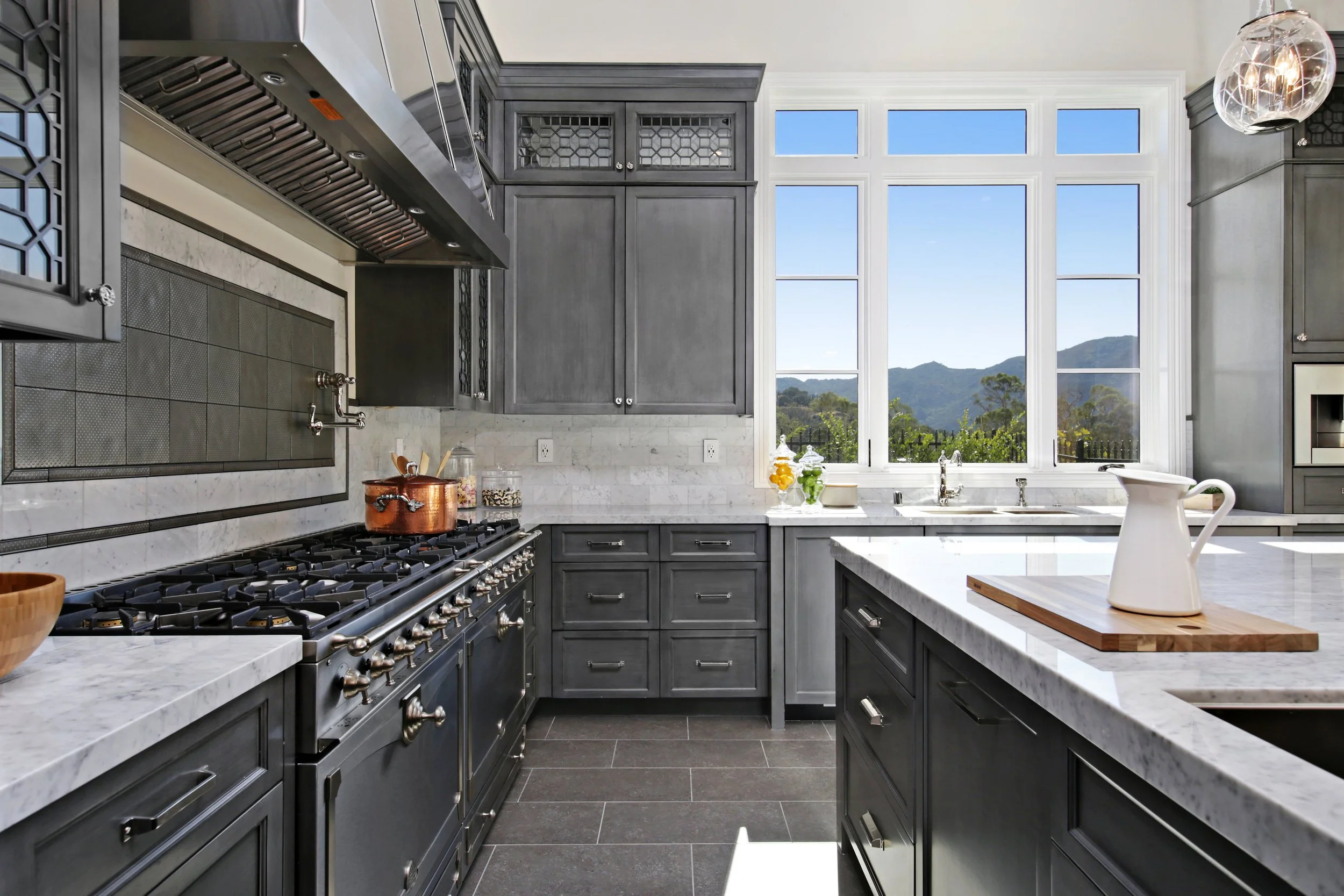 Modern kitchen with gray cabinetry, marble countertops, a large window with mountain view, and kitchen appliances.