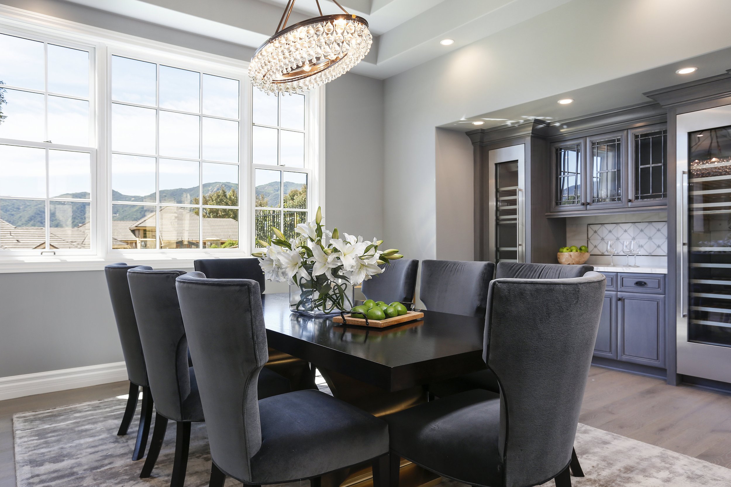 A dining room with a black dining table, six dark gray upholstered chairs, a glass vase with white lilies, and a tray of green apples. Large windows reveal a view of houses and mountains outside. The room has a modern chandelier and built-in gray cab