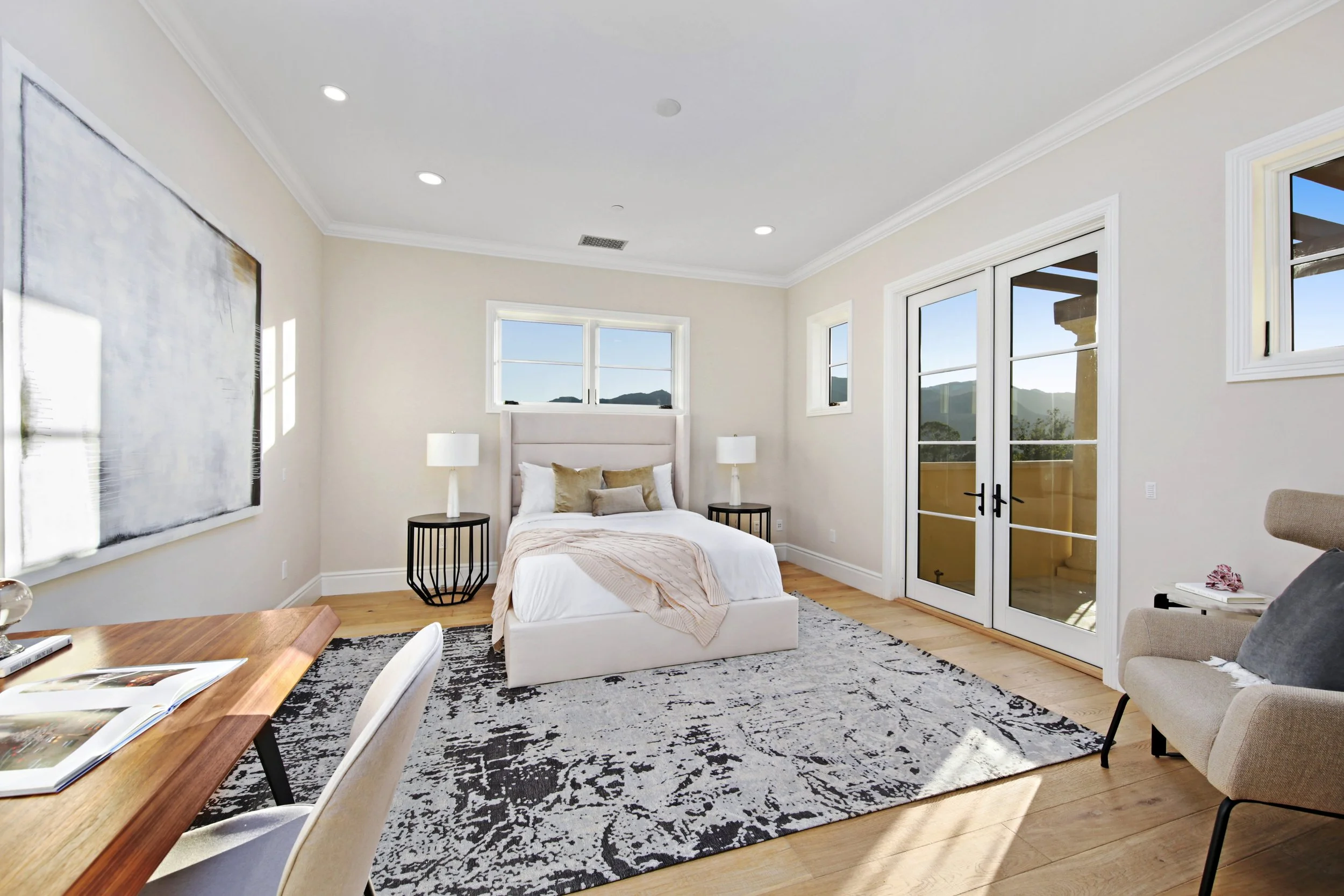Bright bedroom with white walls, wooden floor, a bed with white bedding, two black nightstands with lamps, and a sliding glass door leading to a balcony with mountain views.