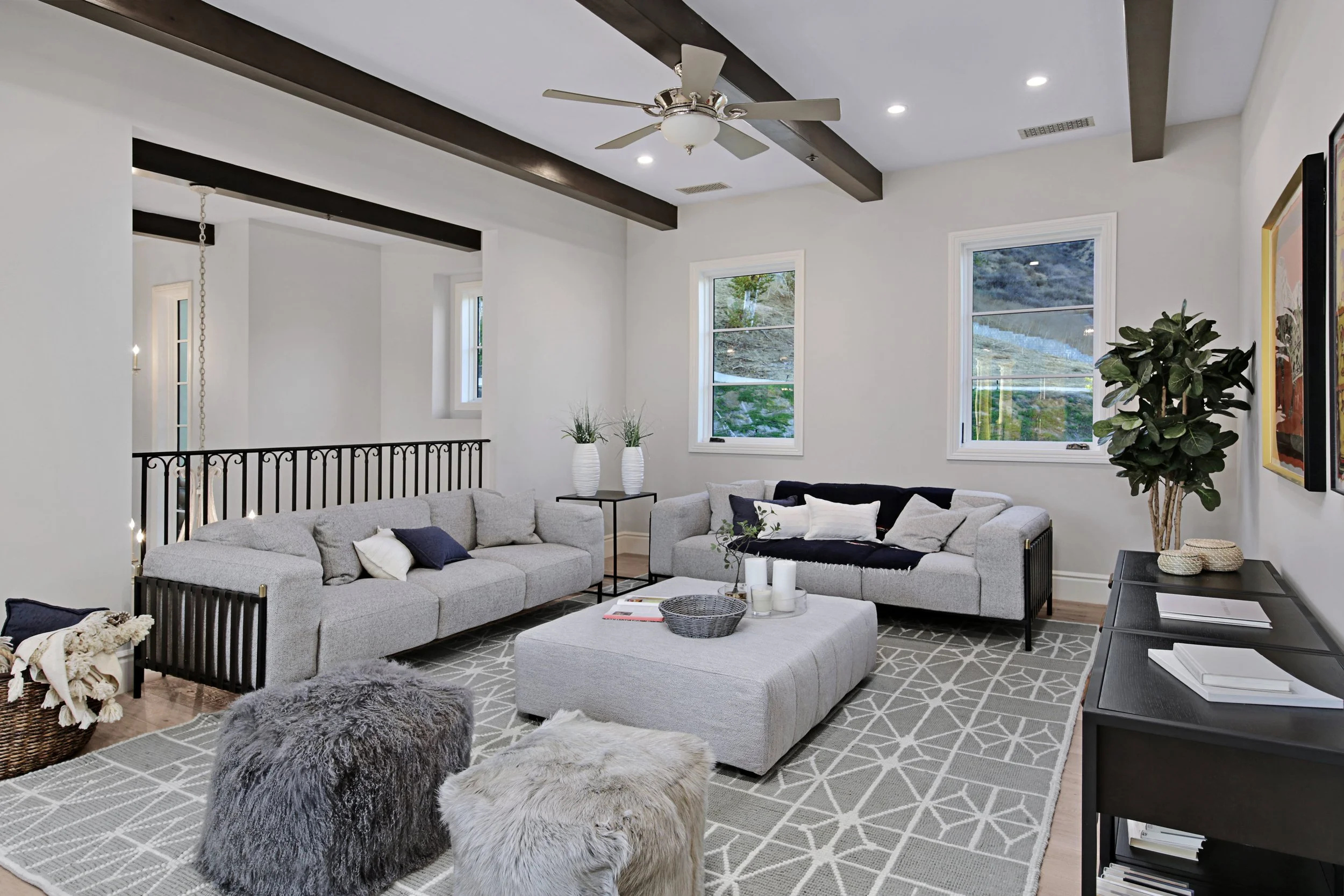 A modern living room with white walls, dark ceiling beams, two large windows, a ceiling fan, two grey sofas adorned with pillows, a rectangular coffee table with candles, a patterned rug, two fluffy ottomans, a black console table with reading materi