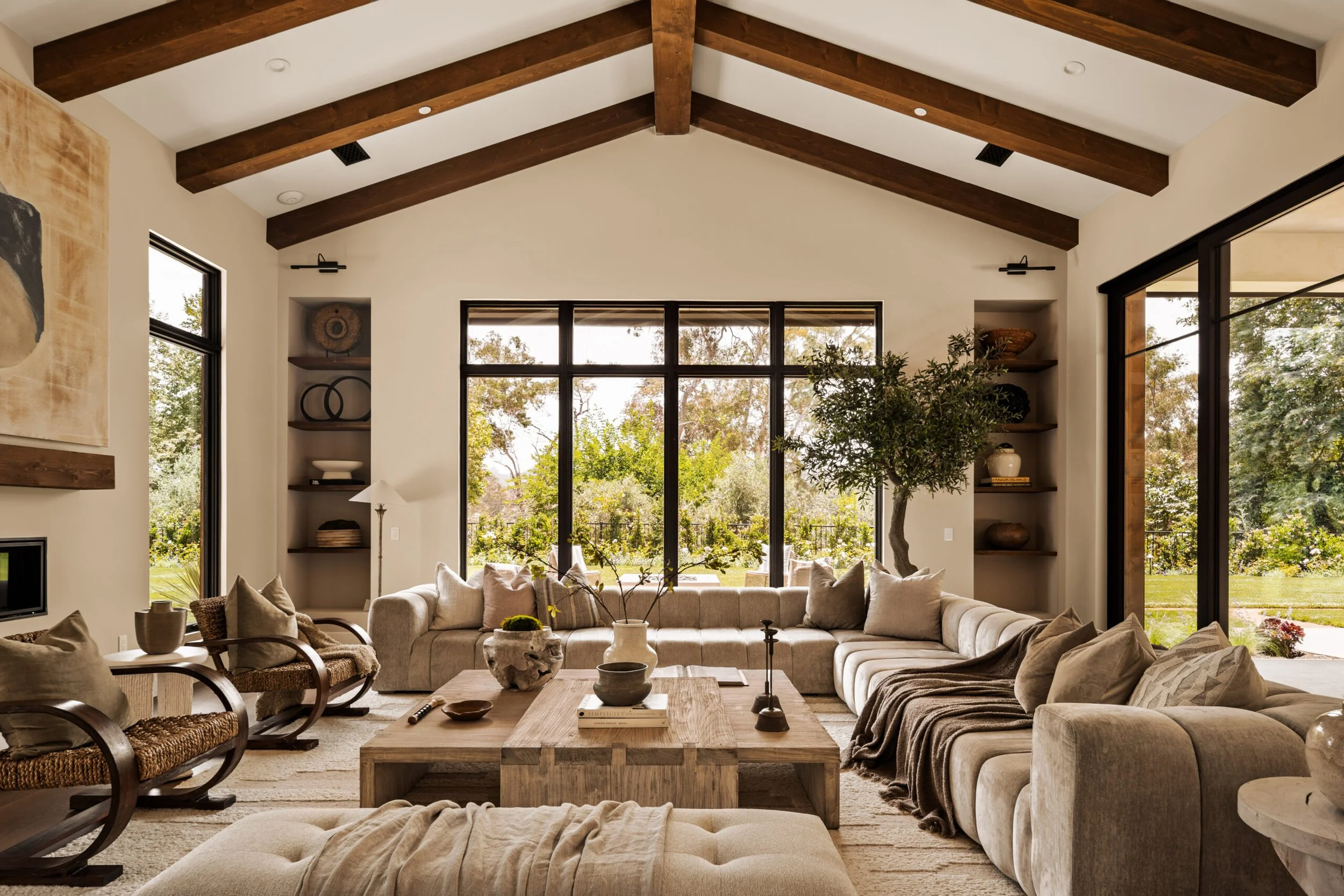 Living room with large beige sectional sofa, wooden coffee table, and armchairs with woven seats, large windows with outdoor greenery, and a tall indoor tree.