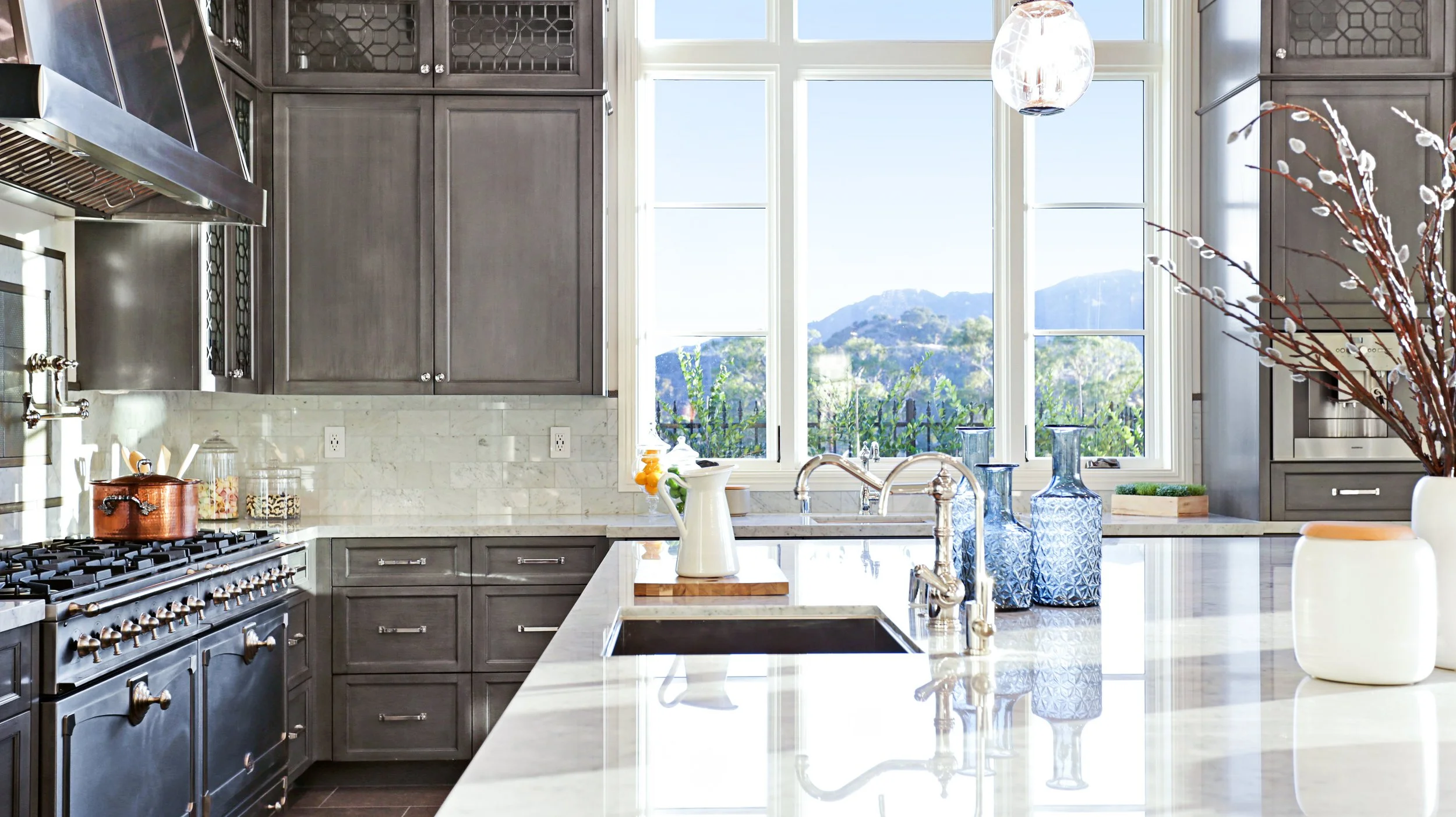 Modern kitchen with gray cabinets, white marble countertops, and a large window showing mountains in the distance. Decor includes blue vases, a white pitcher on a cutting board, and a vase with branches.