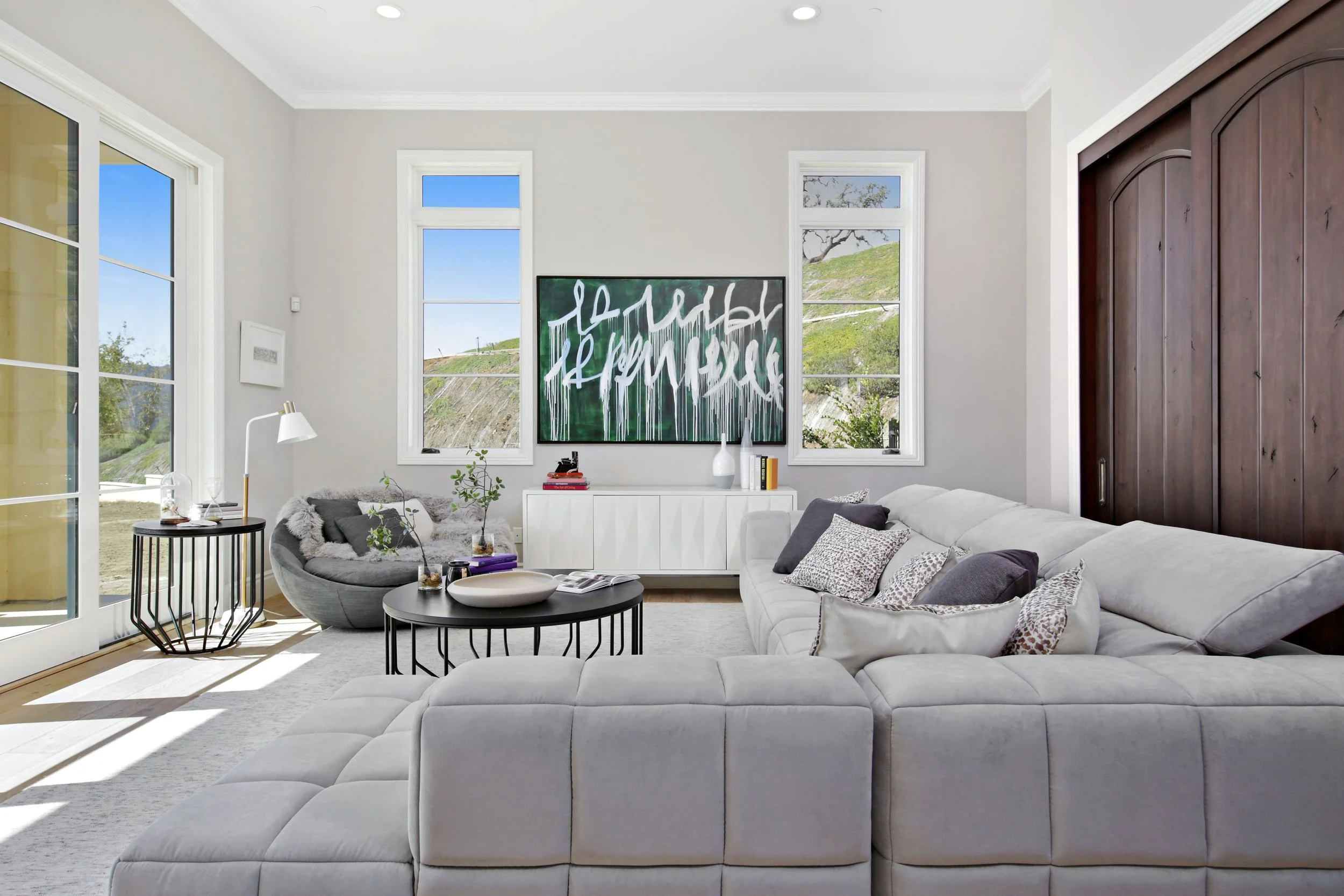 Bright living room with white walls, large windows, white sectional sofa with pillows, round black coffee tables, gray lounge chair with blanket, and a dark wood cabinet.