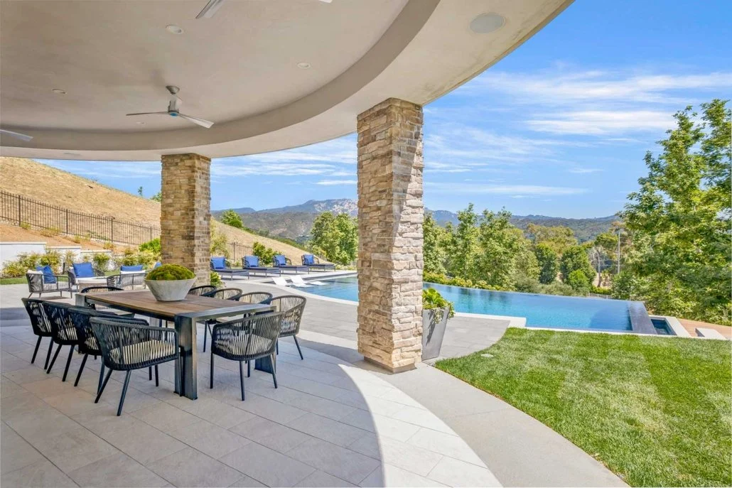 Outdoor patio with dining table, chairs, and a view of a pool, green yard, trees, and distant hills under a blue sky.
