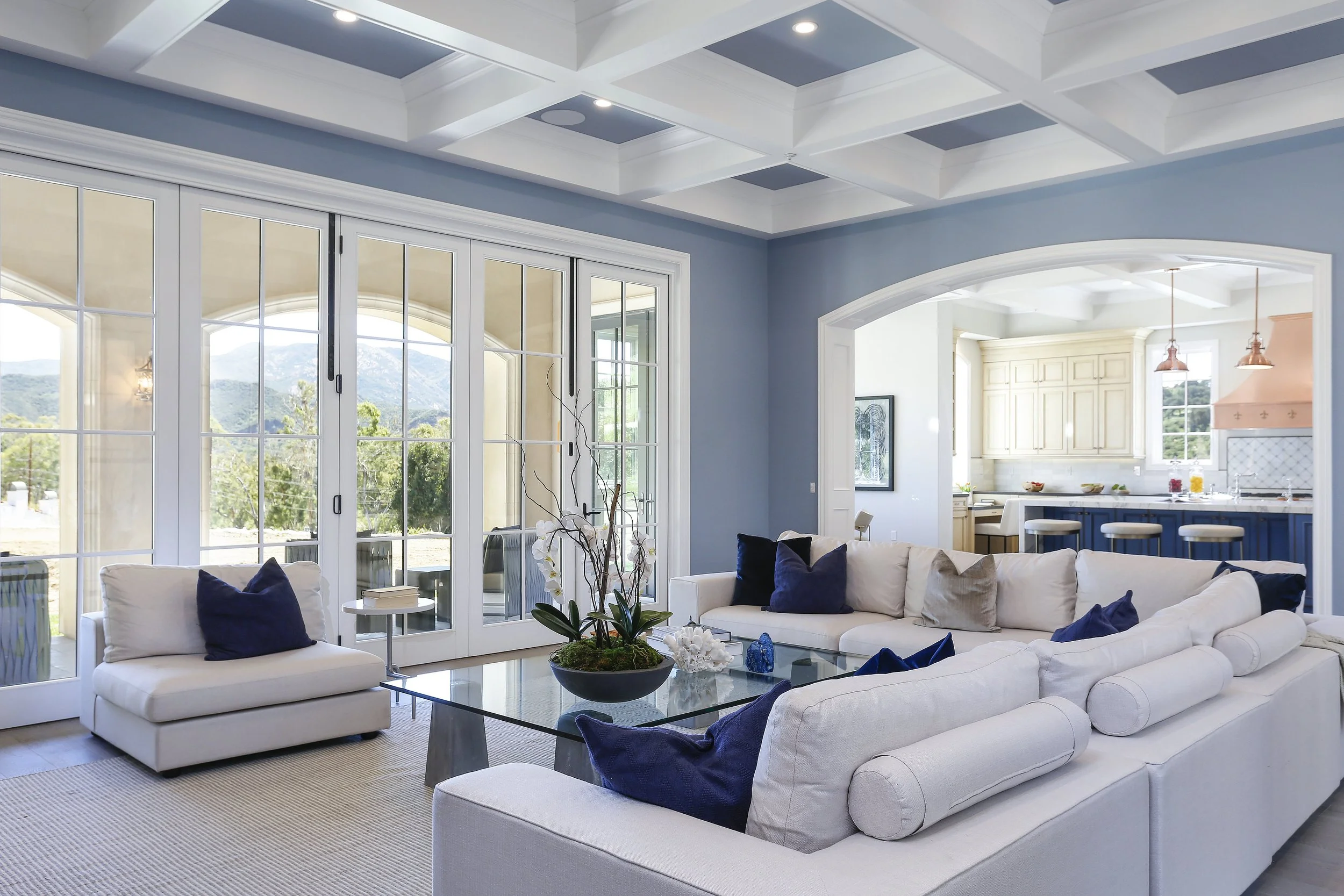 Bright living room with large glass doors, white sectional sofa with navy and beige pillows, glass coffee table with decorative items, view of mountains outside, open kitchen with white cabinets and navy island in the background.