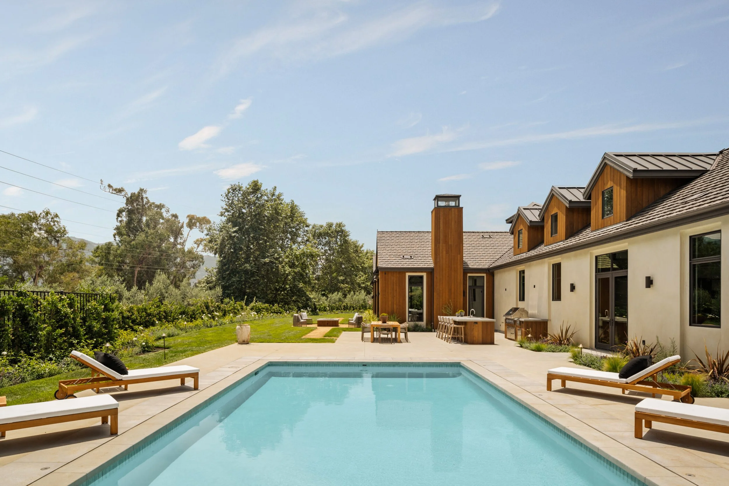 Residential backyard with a swimming pool, lounge chairs, outdoor dining area, built-in grill, and a house with a wooden and stucco exterior, surrounded by trees and greenery.