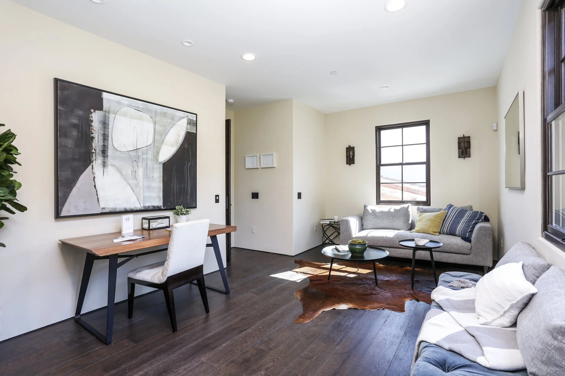 Modern living room with beige walls, dark hardwood floors, a large abstract black and white painting, a light gray sofa with throw pillows, a cowhide rug, and a wooden desk with a white chair.