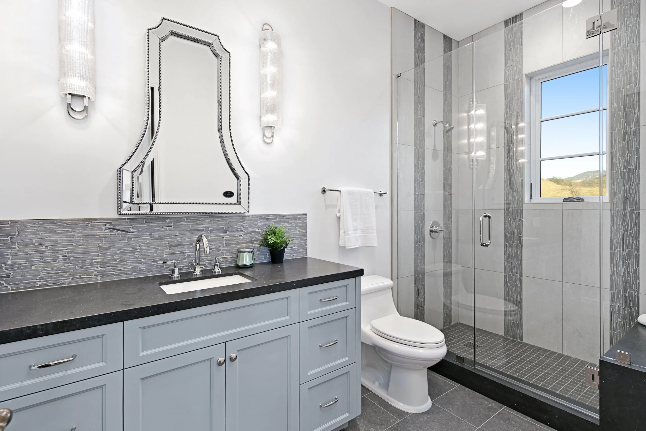 Modern bathroom with a vanity, mirror, wall sconces, toilet, and walk-in shower with window.