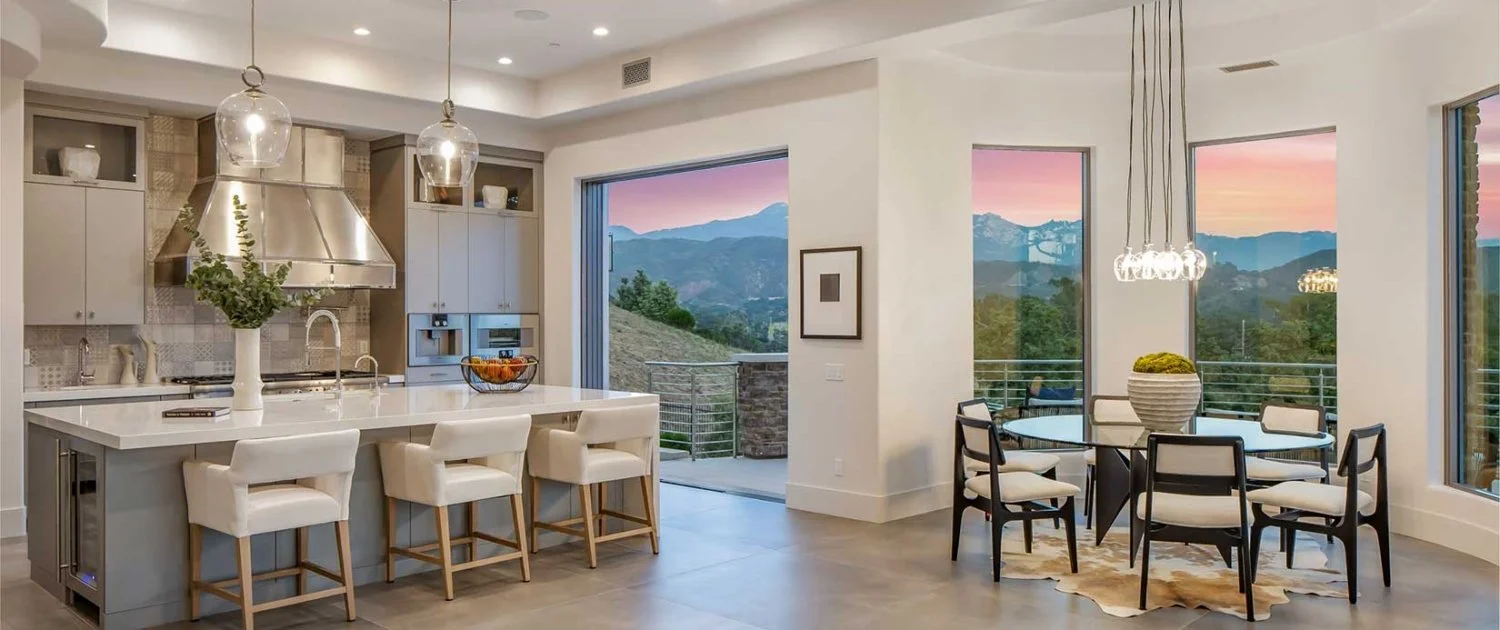 Modern kitchen and dining area with large windows showing a mountain view during sunset.