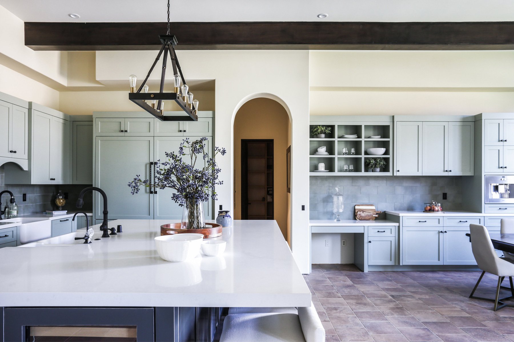 Modern kitchen with light gray cabinets, white countertops, and a central island with a vase of purple flowers. There is a black chandelier hanging from the ceiling and a spacious, organized open shelving unit on the right wall. The floor is tiled in