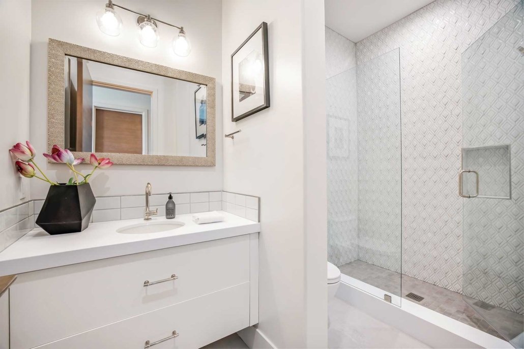 A modern white bathroom with a vanity, large mirror, three light bulbs, pink flowers, framed artwork, and a glass-enclosed walk-in shower with geometric tile pattern.