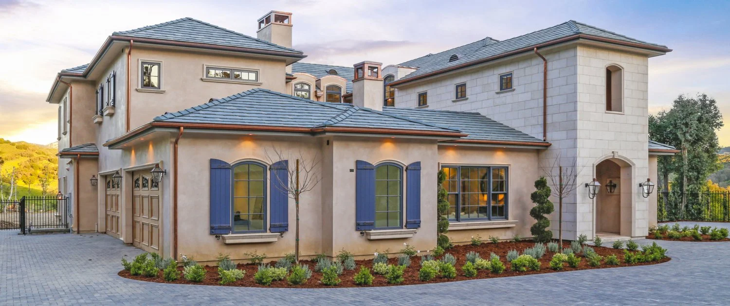 A large, elegant house with stucco walls, blue shutters, and a stone entryway. The property has a paved driveway, landscaped garden, and a black iron fence in the background, with trees and a sunset sky.